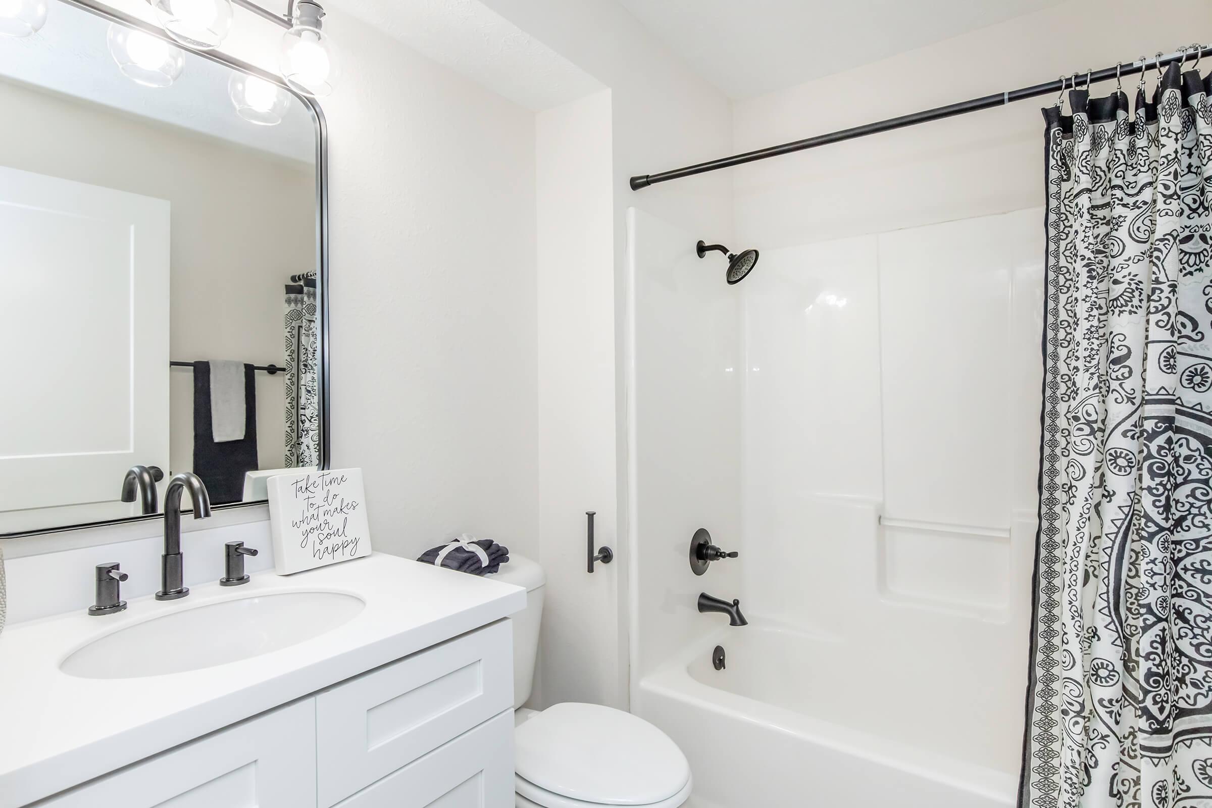 A modern bathroom featuring a white vanity with a mirror, a shower/tub combination with a black and white patterned shower curtain, and a white toilet. The space has neutral-colored walls and a small decorative sign on the countertop. Bright lighting from an overhead fixture enhances the clean and contemporary look.