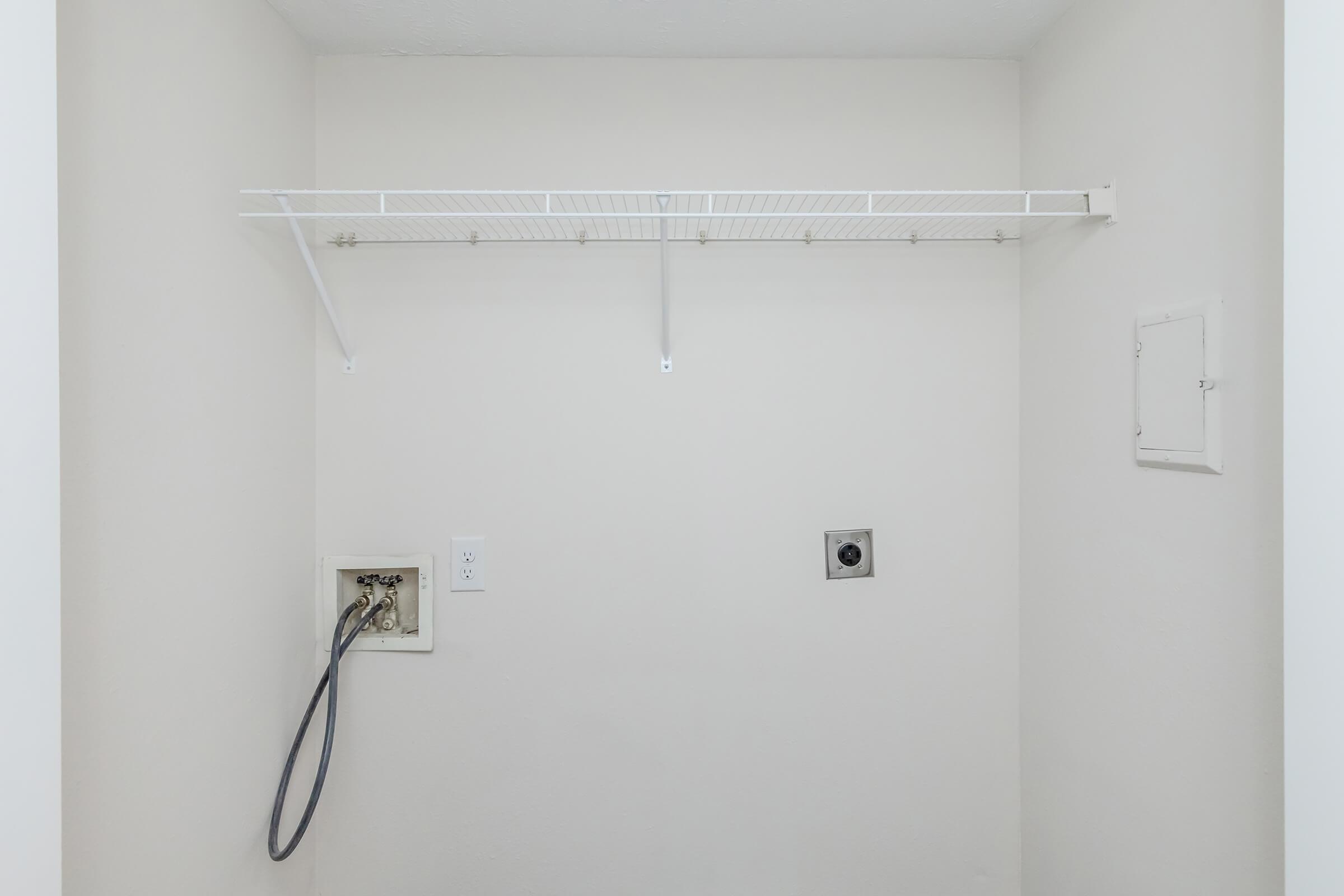 An empty laundry room with a white wall and a metal shelf mounted on the wall. There is a wall outlet and a water supply connection on the left, while a small access panel is visible on the right. The room is uncluttered and well-lit, giving it a spacious appearance.