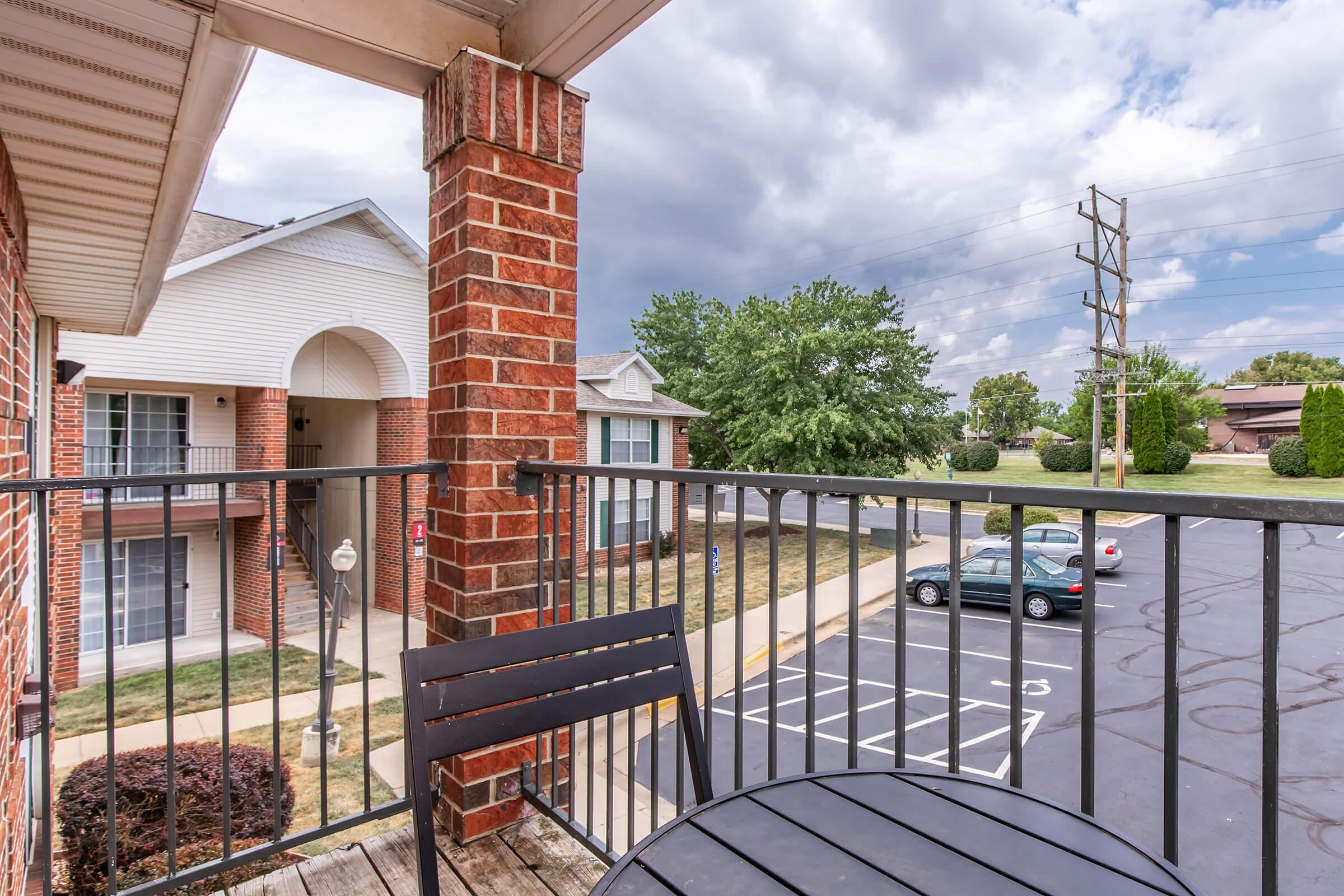 View from a balcony featuring a small round table and chairs, with brick pillars. In the background, there are residential buildings and a parking lot with a couple of parked cars. The sky is partly cloudy, creating a calm atmosphere.