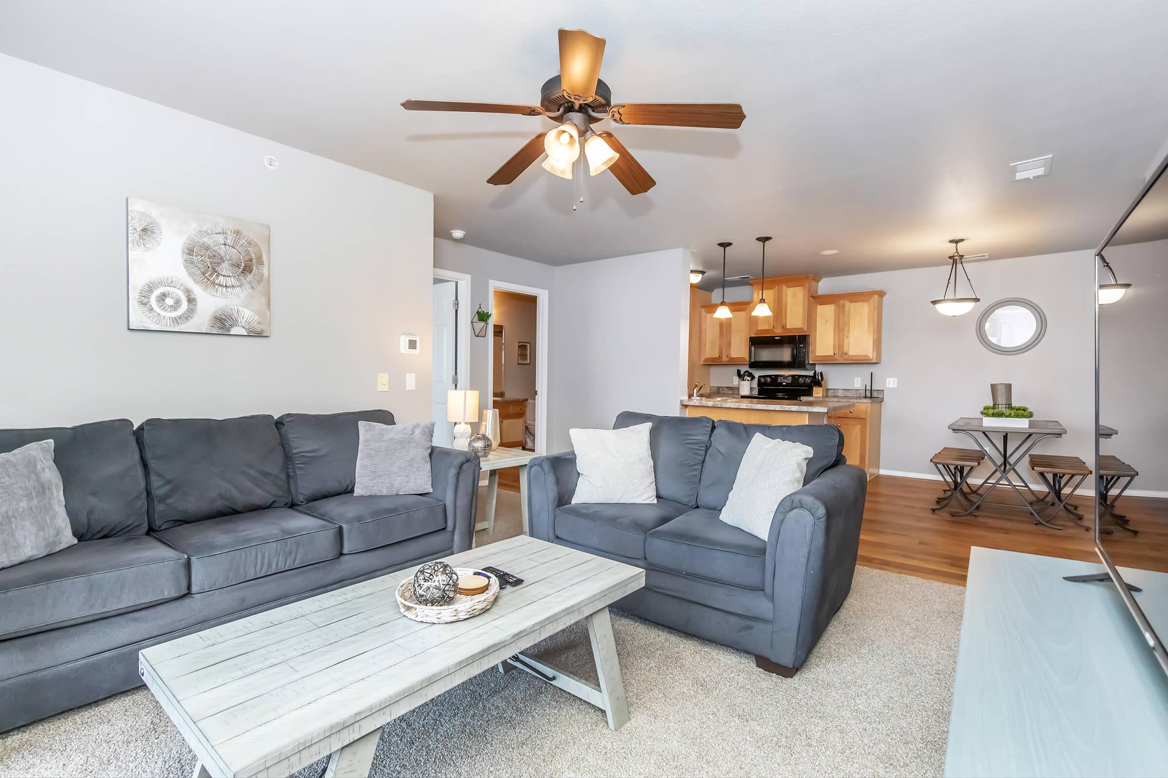 Cozy living room featuring two gray sofas with decorative pillows, a light wooden coffee table with a centerpiece, and a ceiling fan. The room includes an open kitchen area with wooden cabinetry and a dining set visible in the background. Walls are painted in a soft gray tone, creating a warm atmosphere.