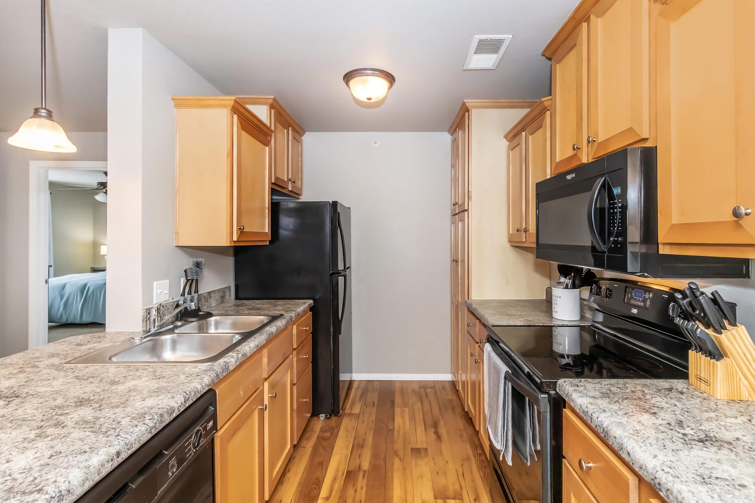 Modern kitchen featuring wooden cabinets, granite countertops, and stainless steel appliances. The layout includes a double sink, a black refrigerator, microwave, and stove, with hardwood flooring. Soft lighting from overhead fixtures adds warmth to the space.