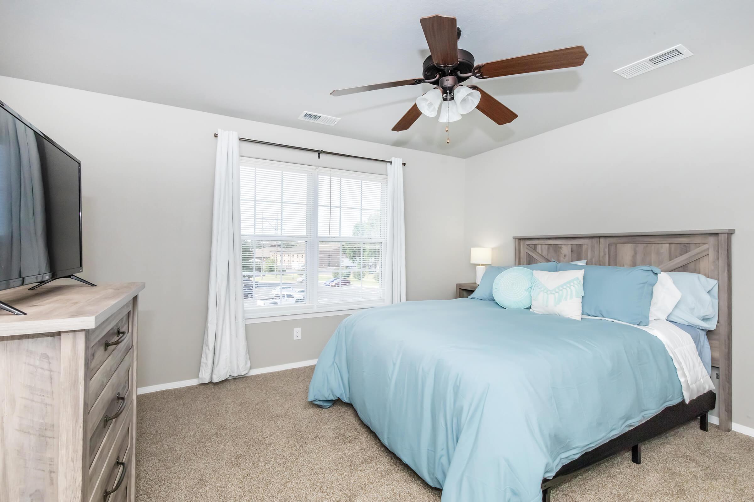 A modern bedroom featuring a queen-sized bed with light blue bedding, a wooden headboard, and a ceiling fan. There's a dresser with a flat-screen TV, and large windows with white curtains that allow natural light in, adding to the cozy atmosphere. The carpet is beige, complementing the neutral wall color.