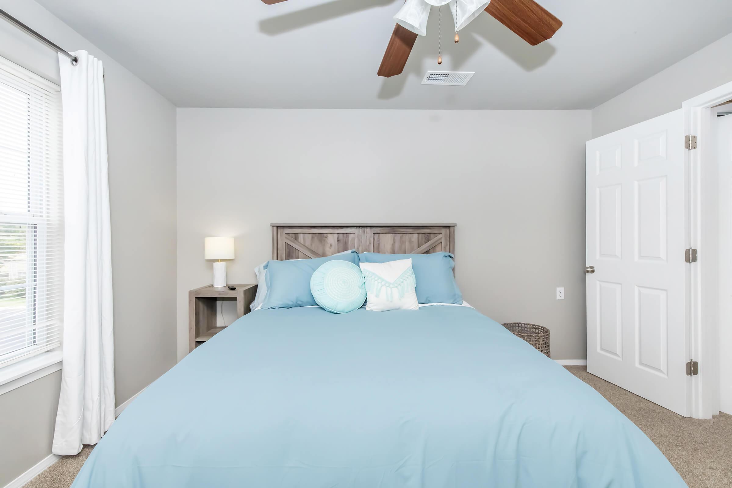 A cozy bedroom featuring a light blue bedspread, decorative pillows, and a wooden headboard. There is a bedside table with a lamp next to the bed, a window with white curtains, and a door leading to another room. The carpeted floor adds warmth to the space.