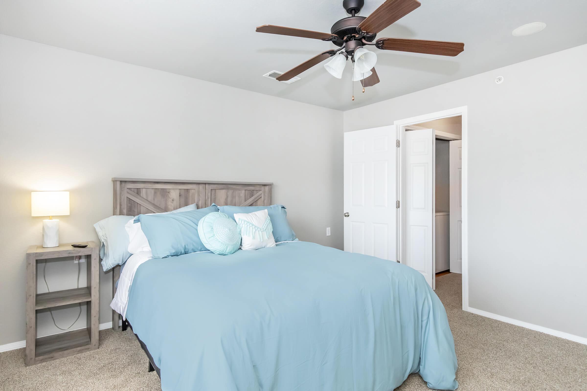 A clean and modern bedroom featuring a light blue bedspread with matching pillows on a queen-sized bed. A wooden nightstand with a lamp stands beside the bed. The room has light gray walls, a ceiling fan, and a doorway leading to another room, creating a bright and inviting atmosphere.