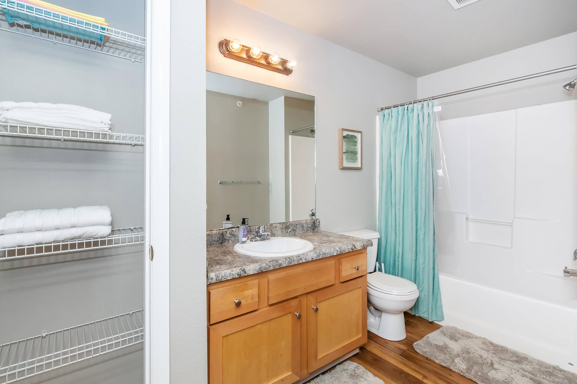 A clean bathroom featuring a large mirror above a granite countertop with a sink, wooden cabinetry, and a white toilet. A shower/tub combo is visible with a light teal curtain. Shelves hold neatly folded white towels. The flooring is a warm wood tone, adding a cozy feel to the space.