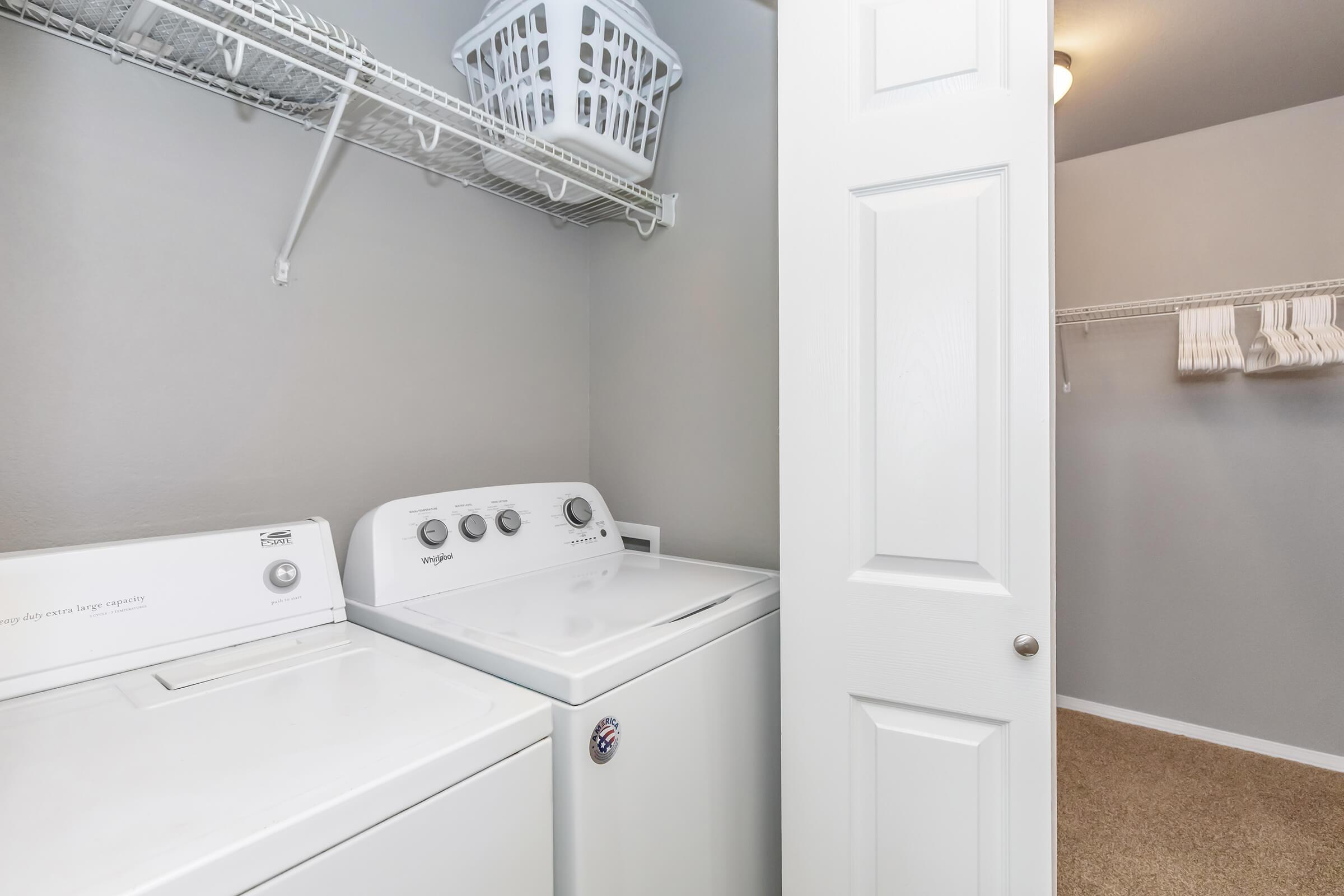 A laundry room featuring a white washing machine next to a white dryer. There is a shelf above with a laundry basket and a door partially open to a closet area with neatly hung towels. The walls are painted light gray, and the floor is covered with carpet.