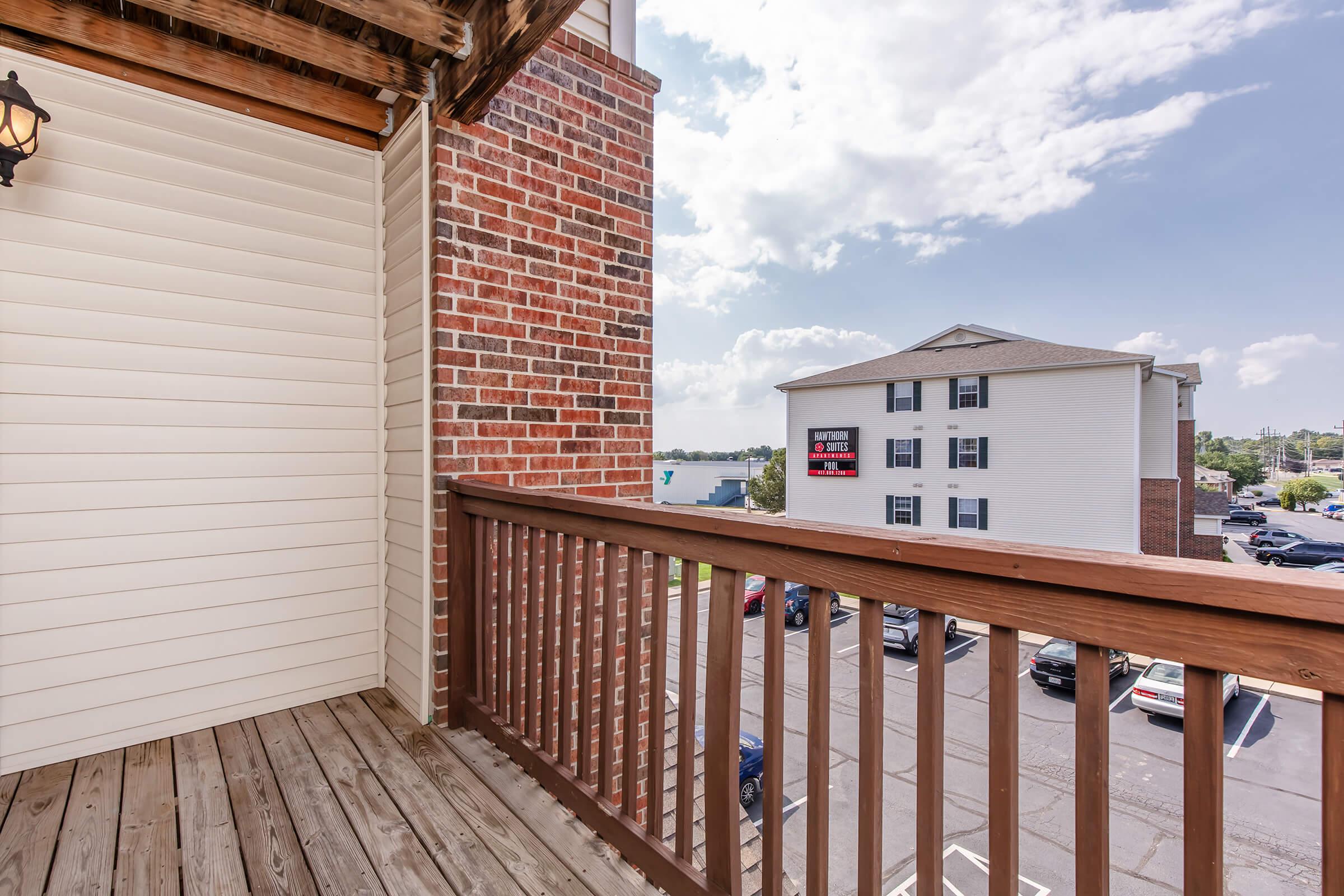 A view from a wooden balcony overlooking a parking lot and surrounding buildings. The sky is partly cloudy with blue patches visible. The balcony features wooden railing, and a brick wall is visible on one side. An apartment building can be seen in the distance with a sign.
