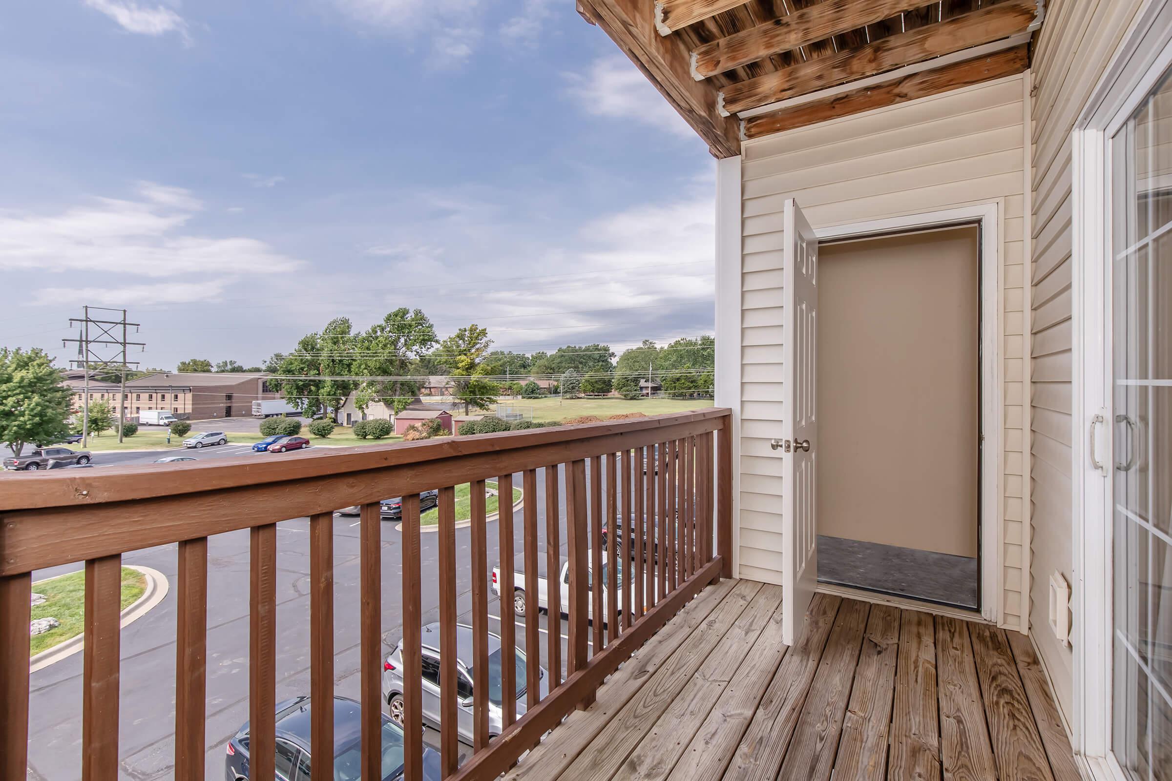 View of a balcony with wooden railing and flooring, showcasing an open door leading into a room. The scene includes a parking area and trees in the distance, under a partly cloudy sky. The area appears well-maintained and residential.