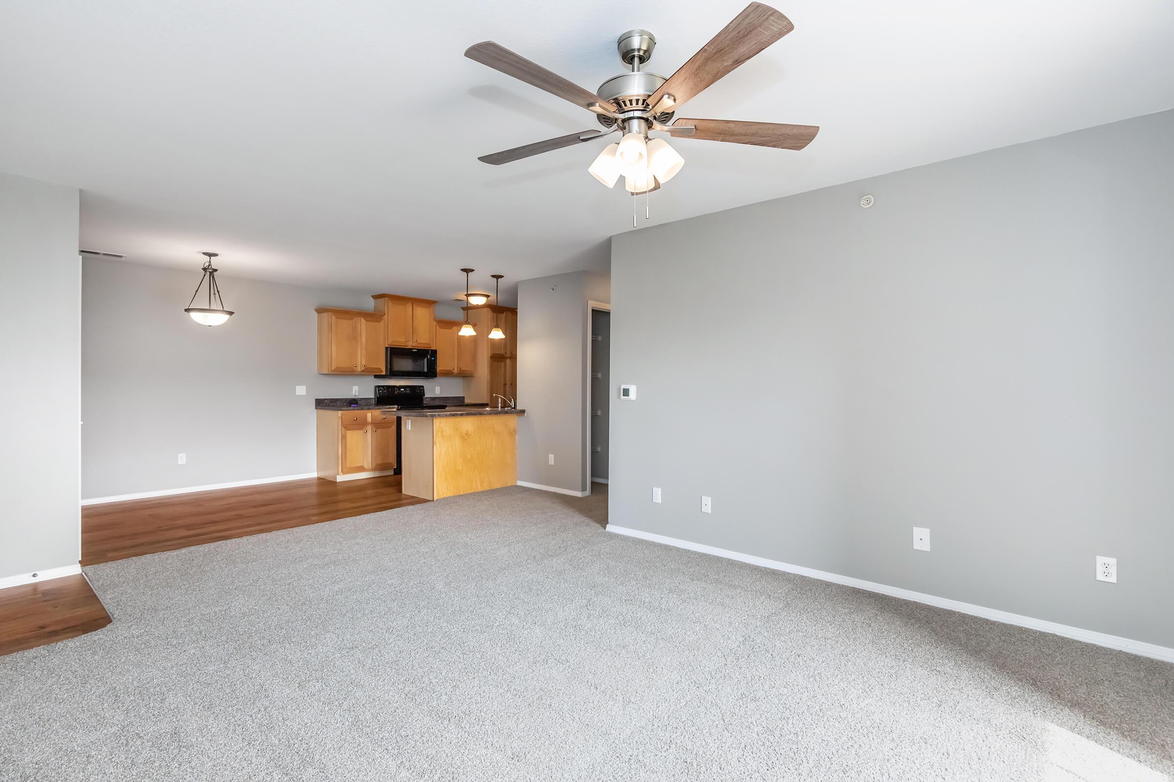 Bright and airy living space featuring light gray walls, a ceiling fan with lights, and a cozy carpeted floor. The open floor plan connects to a kitchen with wooden cabinetry and black appliances. Soft lighting fixtures enhance the welcoming atmosphere. Ideal for comfortable living or entertaining.
