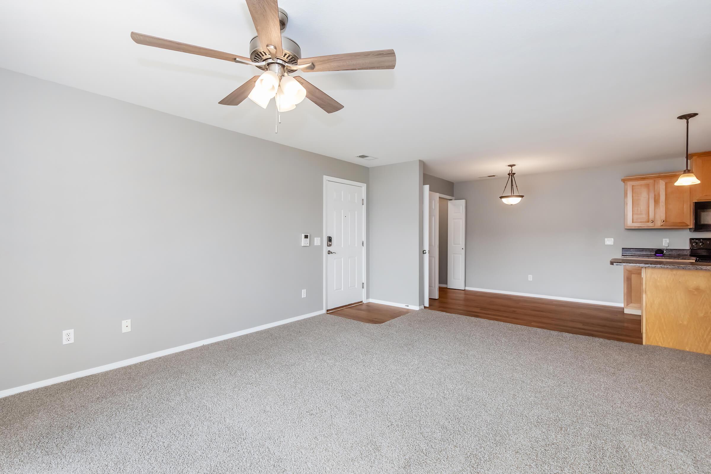 Spacious living room with light gray walls and a ceiling fan. Featuring a plush carpeted floor and an open doorway leading to other areas. A kitchen area is visible in the background with wooden cabinetry and lighting fixtures. Natural light fills the space, creating a warm and inviting atmosphere.