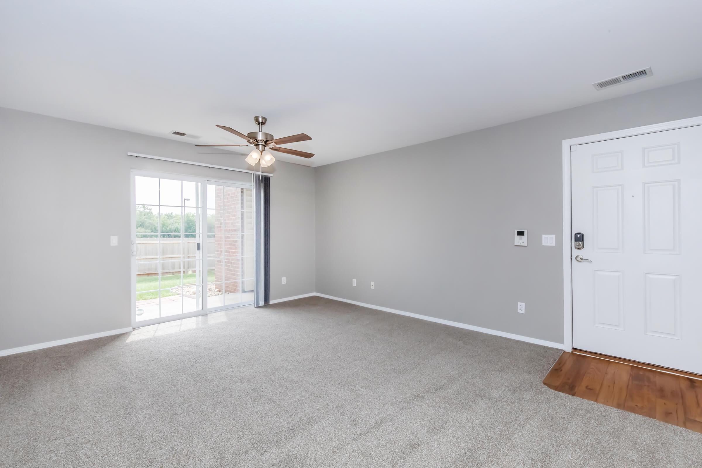 An empty living room featuring light gray walls, a ceiling fan, and beige carpet. A sliding glass door leads to an outdoor area, providing natural light. The front door is visible on the right, and there is a small panel by the door, likely for a security system or intercom.