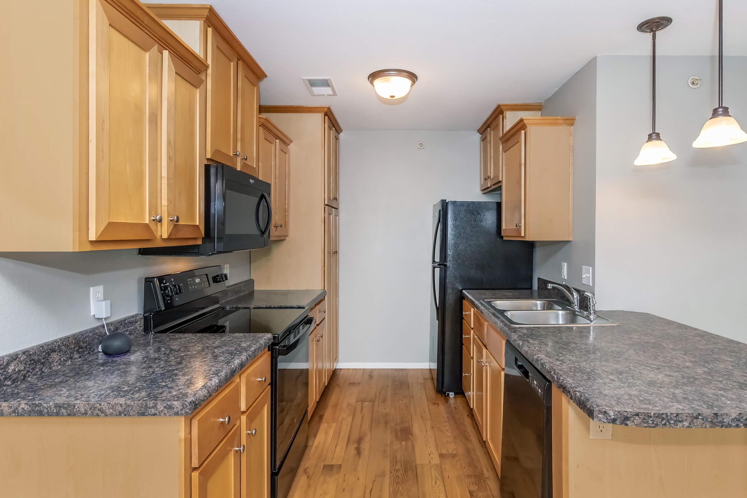 Modern kitchen with wooden cabinets, granite countertops, a black microwave, oven, and refrigerator, along with a stainless steel sink. Pendant lights are hanging above the counter, and the flooring is wooden. The overall design is clean and contemporary, emphasizing functionality and style.