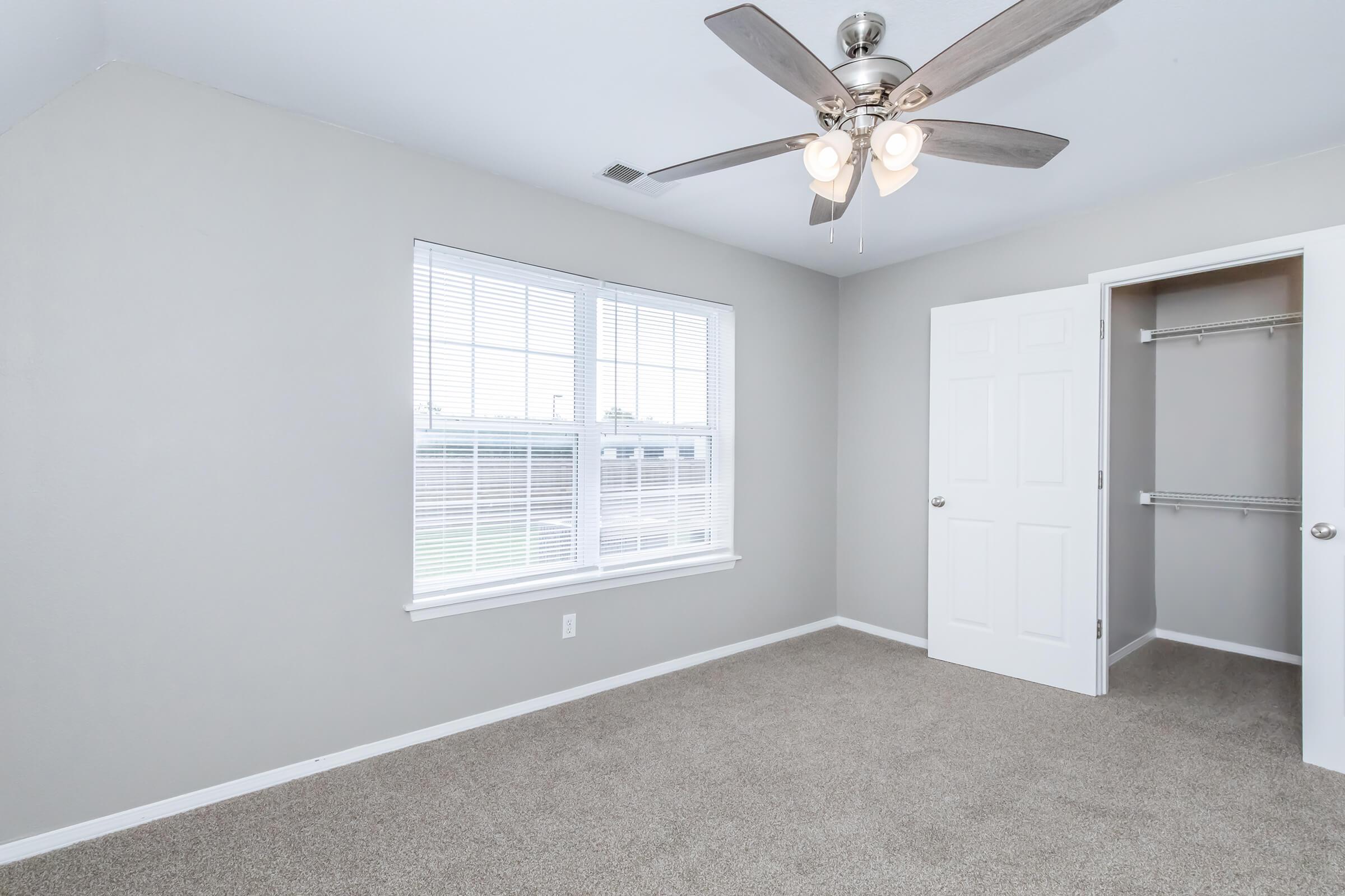 A spacious, light gray bedroom featuring a ceiling fan with light fixtures, a large window with white blinds, and a closet with a white door. The floor is carpeted in a neutral tone, creating a clean and inviting atmosphere.