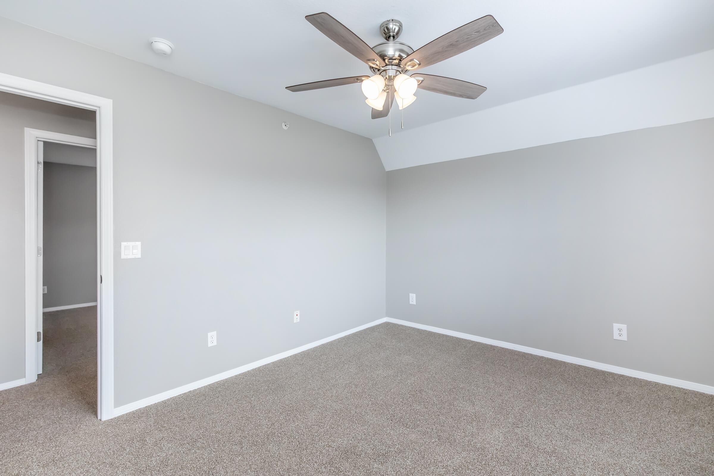 Empty room with light gray walls and carpeted floor. A ceiling fan with four lights hangs from the ceiling, and there are doors leading to other rooms visible in the background. The overall atmosphere is bright and open, suitable for various purposes.