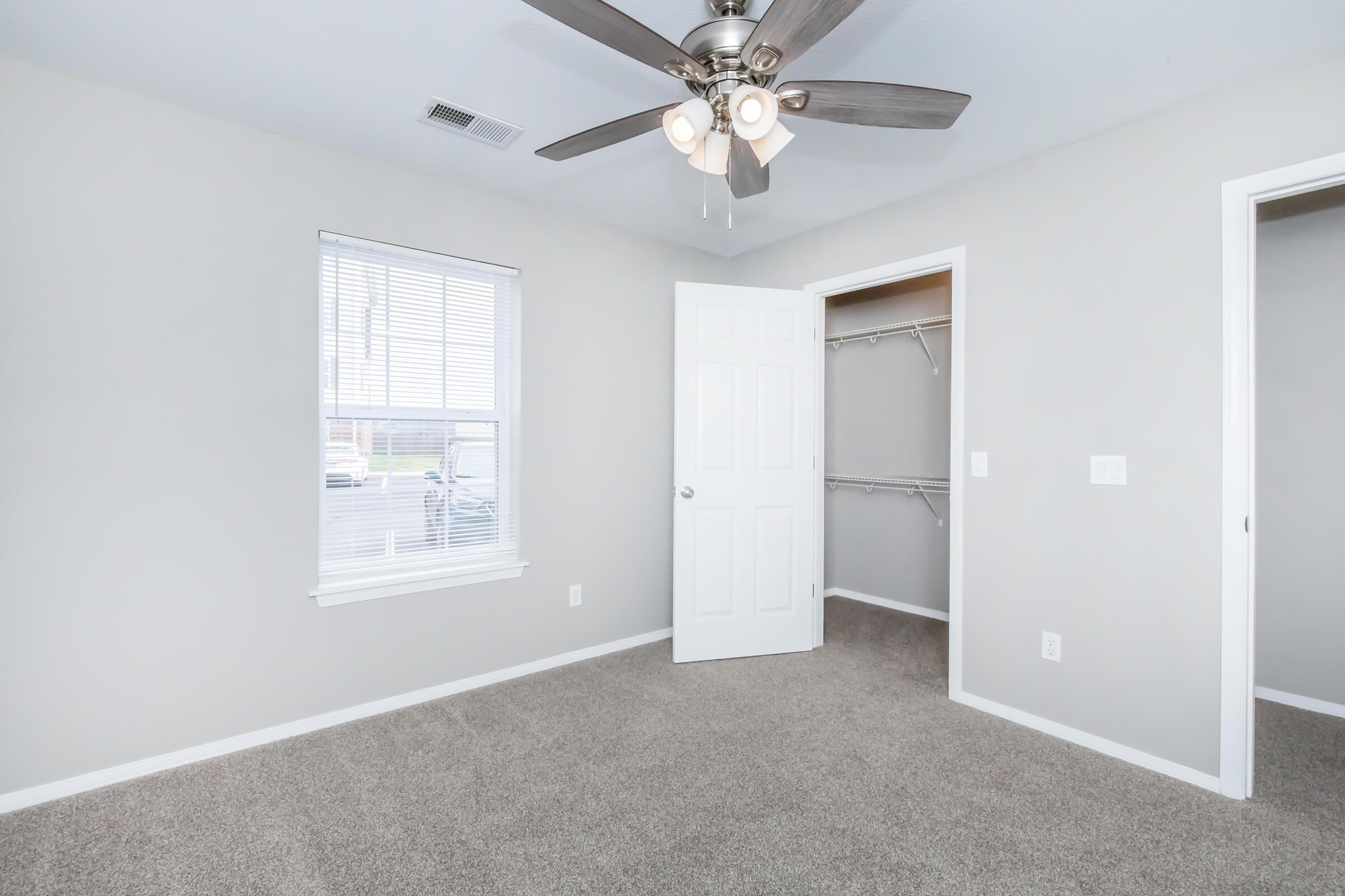 A well-lit bedroom featuring light gray walls, a ceiling fan with three bulbs, and beige carpeting. There are two doorways, one leading to a closet with shelving and the other to another room. A window provides natural light, and the overall space appears clean and inviting.