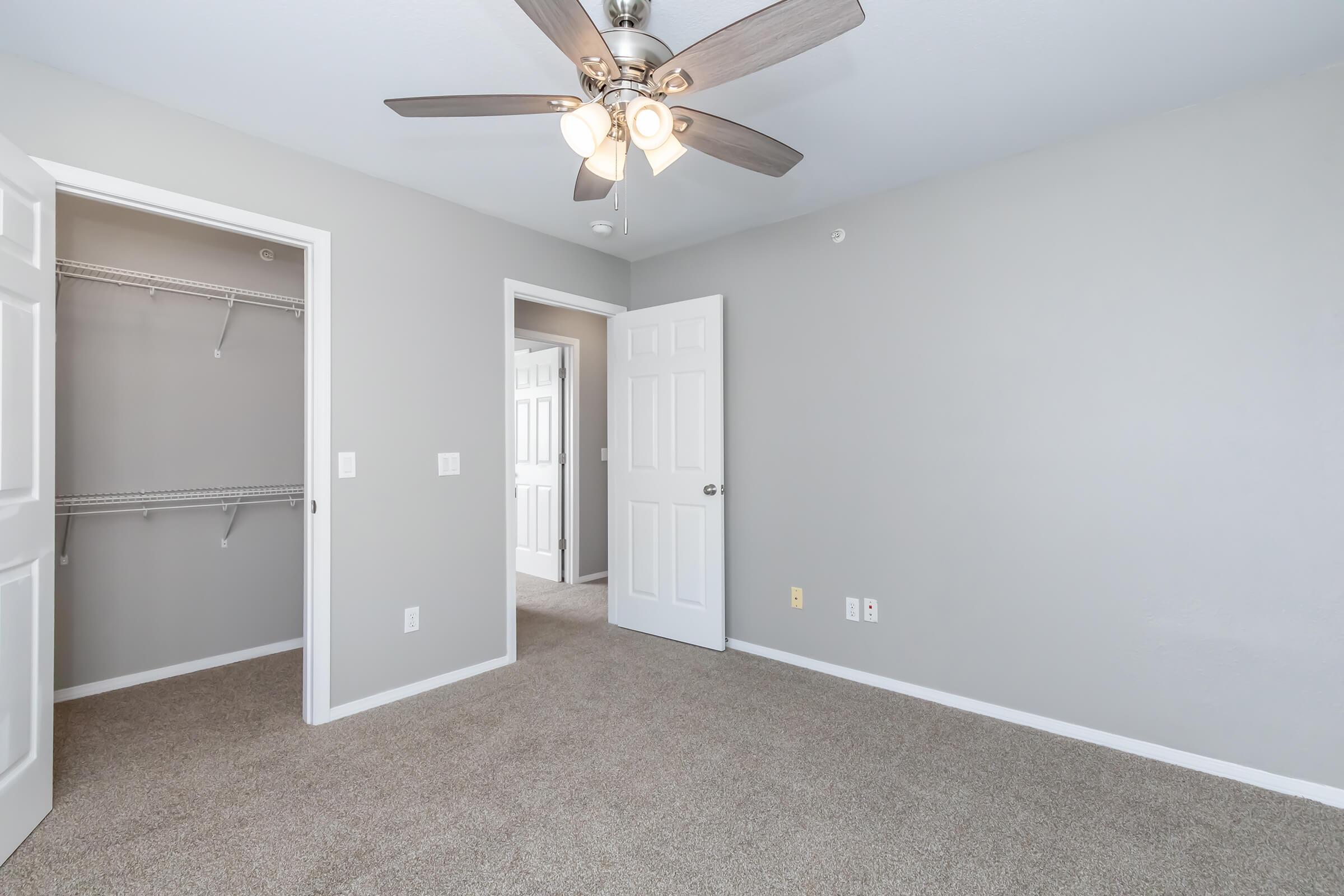 A light-colored bedroom featuring a ceiling fan with wooden blades. There are two open doors leading to a closet and an adjacent room. The walls are painted gray, and the floor is covered with beige carpet. Natural light brightens the space, creating a cozy atmosphere.