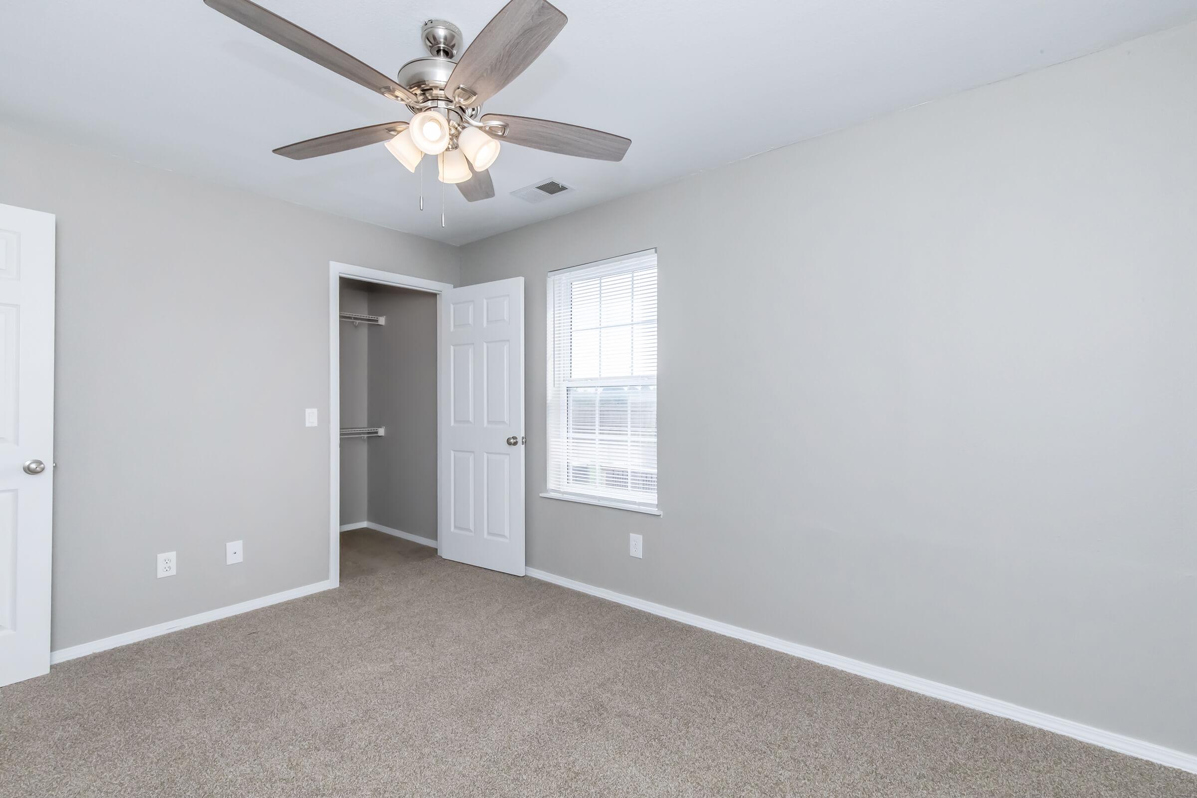 Empty room with light gray walls and a ceiling fan. The floor is covered in beige carpet, and there is a closet with an open door to the left. A window with white blinds allows natural light to enter the space. The room has two doors: one leads to the closet, and the other to another area.