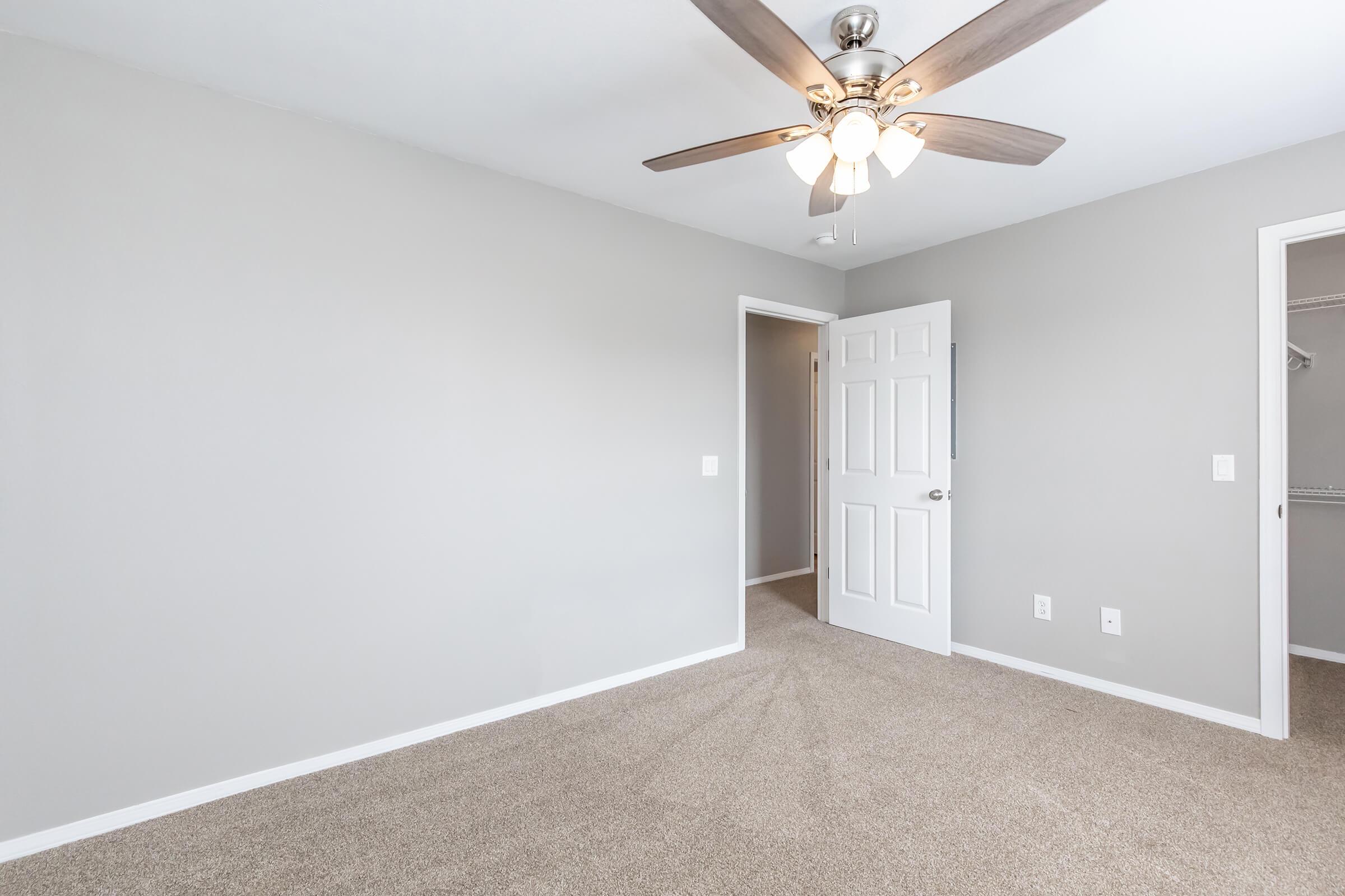 A well-lit, empty bedroom featuring gray walls and beige carpet. The ceiling has a modern ceiling fan with light. One door leads to a closet, and another door opens to a corridor. The room has a minimalist design, creating a spacious and inviting atmosphere.