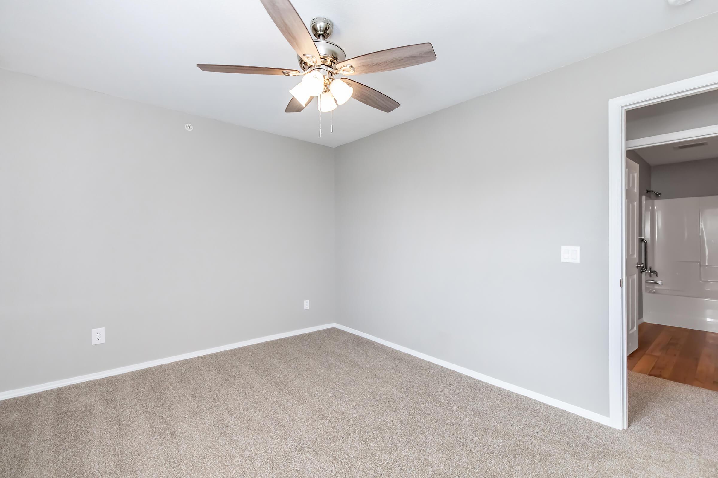 A vacant room with light gray walls and a ceiling fan with wooden blades. The carpet is light beige and the space is well-lit. A doorway on the right leads to a bathroom, which features a shower and tub combination.