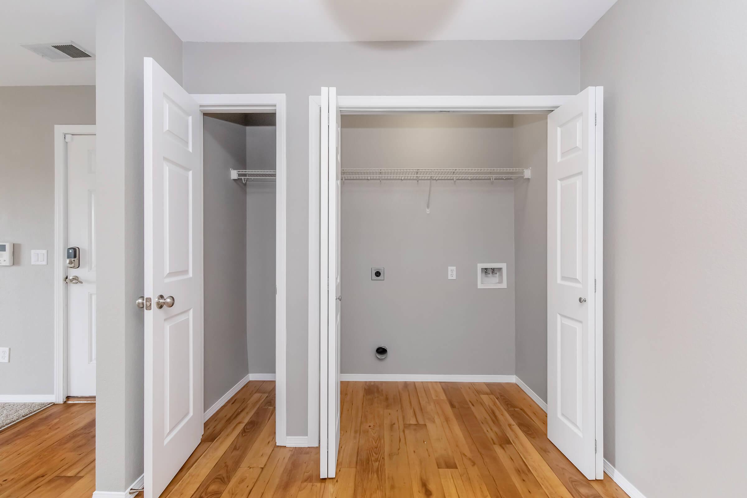 A bright room featuring two open closet doors revealing empty spaces with wooden flooring. The walls are painted a light gray, and there is a small utility area visible in one closet. The room's overall design is clean and modern, emphasizing ample storage potential.