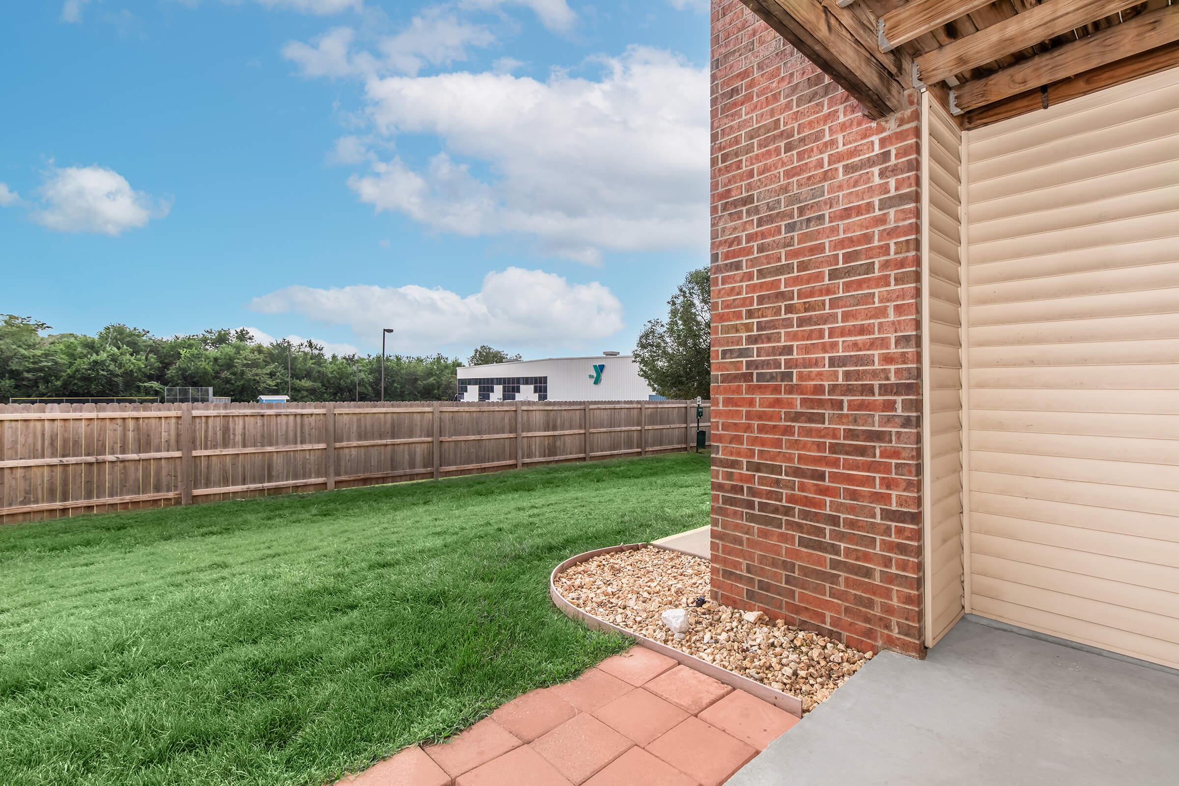 A view of a backyard with green grass and a stone border. There is a wooden fence surrounding the area, and a brick wall on one side. In the background, a building is visible under a blue sky with clouds. The patio area features concrete flooring and a landscaped edge.