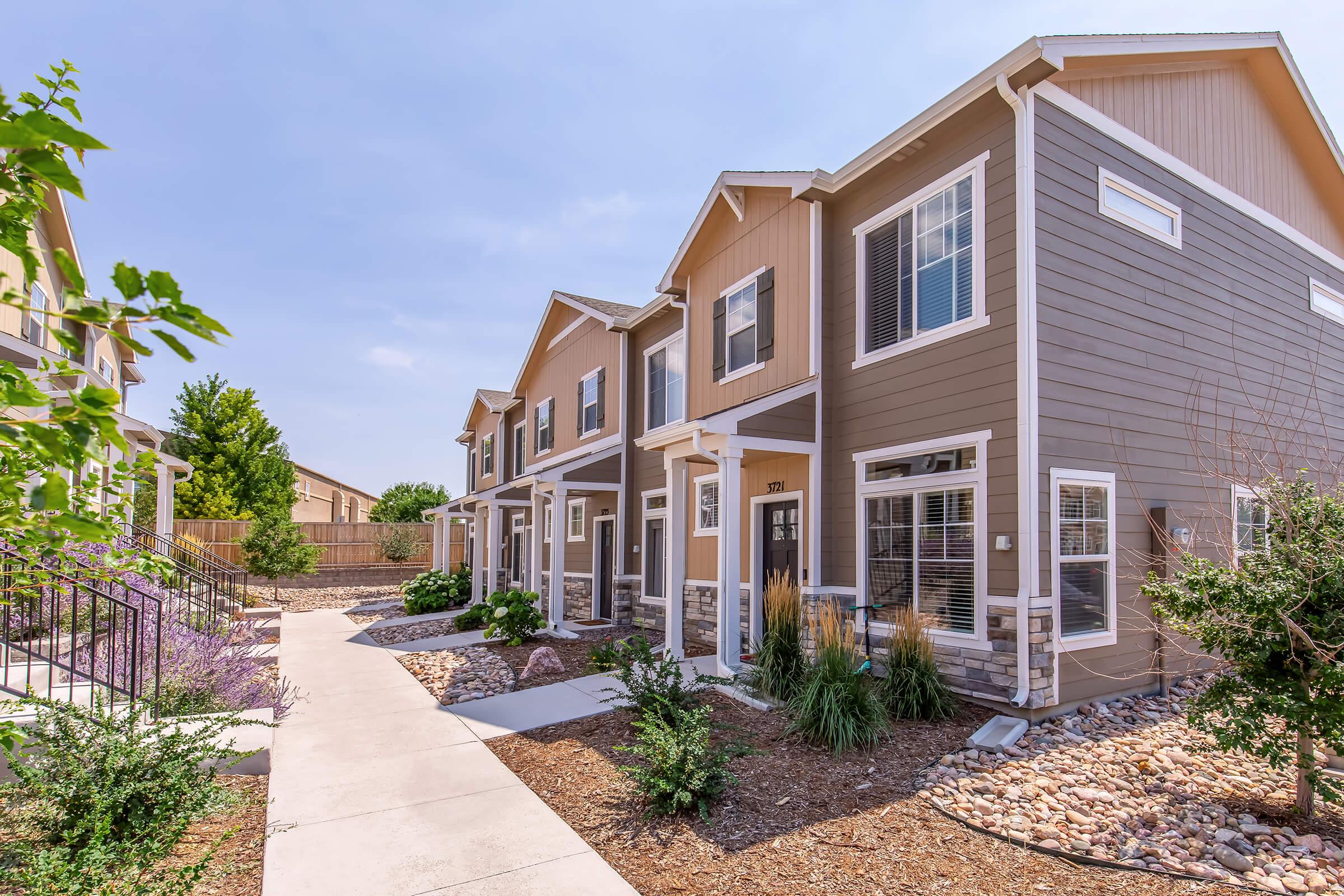 Row of modern townhouses featuring light and dark exterior contrasts, surrounded by landscaped gardens and pathways. Clear blue sky overhead, with green trees and colorful plants enhancing the inviting ambiance of the neighborhood. Ideal for showcasing residential living spaces in a community setting.