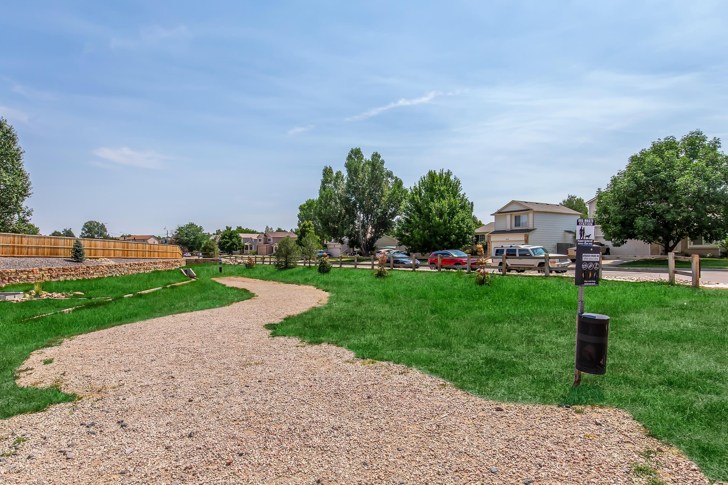 A winding gravel path through a grassy area leads to a nearby residential neighborhood. The scene features trees, a mailbox, and parked cars along the street, under a blue sky with some clouds. The landscape is well-maintained, creating a serene suburban atmosphere.