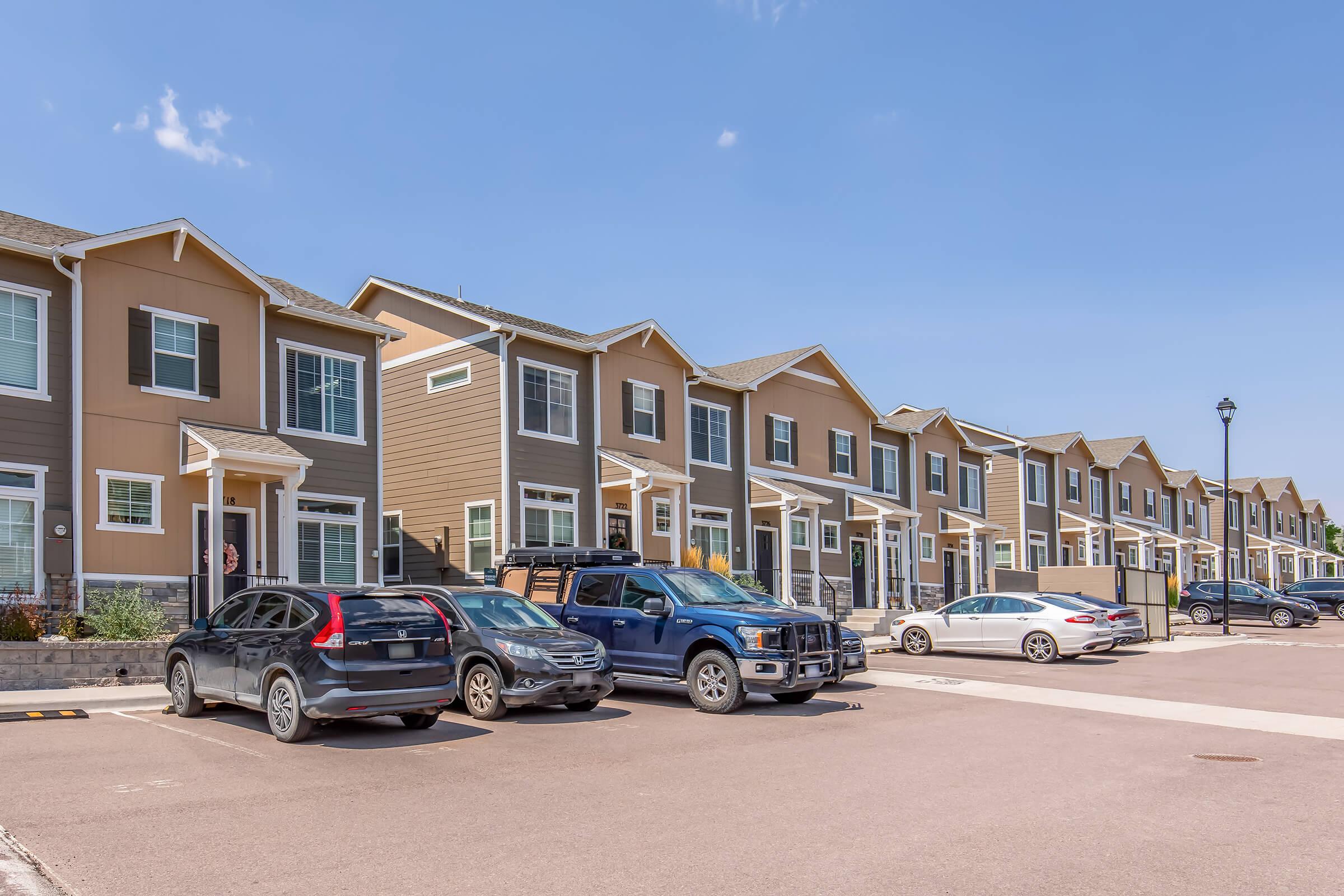 Row of modern two-story townhomes in a suburban area, featuring a variety of exterior colors. Several parked vehicles in the foreground, including SUVs and a pickup truck. Clear blue sky above, with a well-maintained street and landscaping.