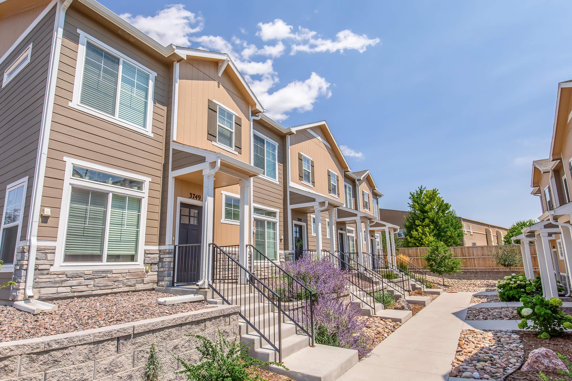 Row of modern townhouses with light brown siding and stone accents, featuring multiple windows and black metal railings. Each unit has a small landscaped area with shrubs and flowers, and the scene is set against a clear blue sky with a few clouds.