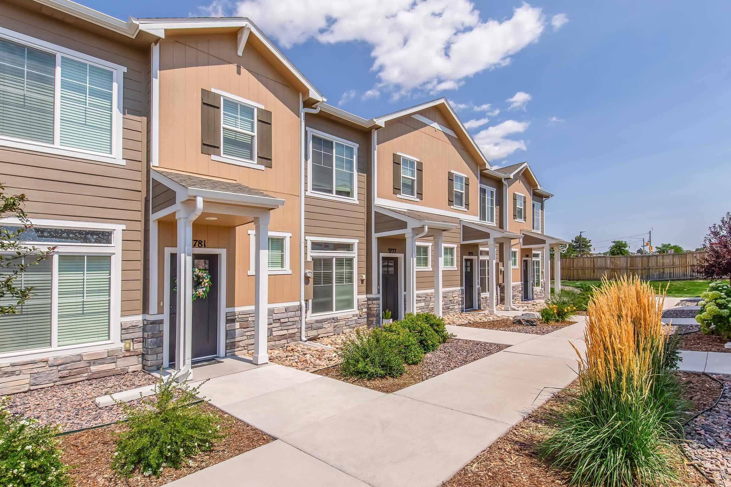 A view of a modern multi-unit residential building featuring several townhouses with brown and beige exteriors. The front porches are shaded, and there is a well-maintained pathway leading to the doors. Lush greenery and decorative landscaping enhance the curb appeal under a sunny sky.
