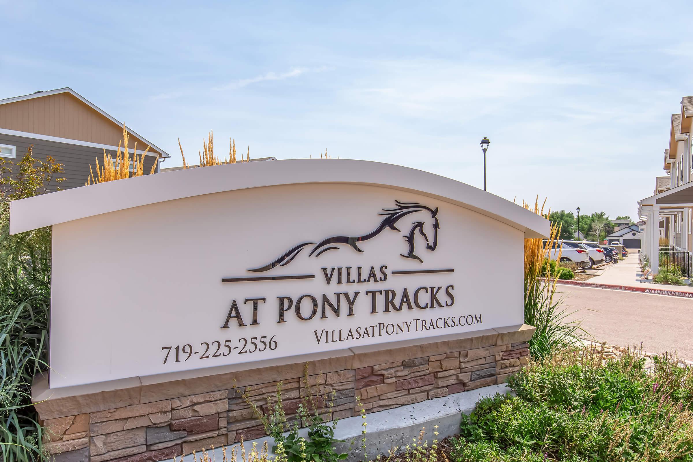 Sign for "Villas at Pony Tracks" featuring a horse silhouette, with contact information and website. Surrounded by landscaped greenery and a sunny sky, it's positioned near residential buildings and a parking area.