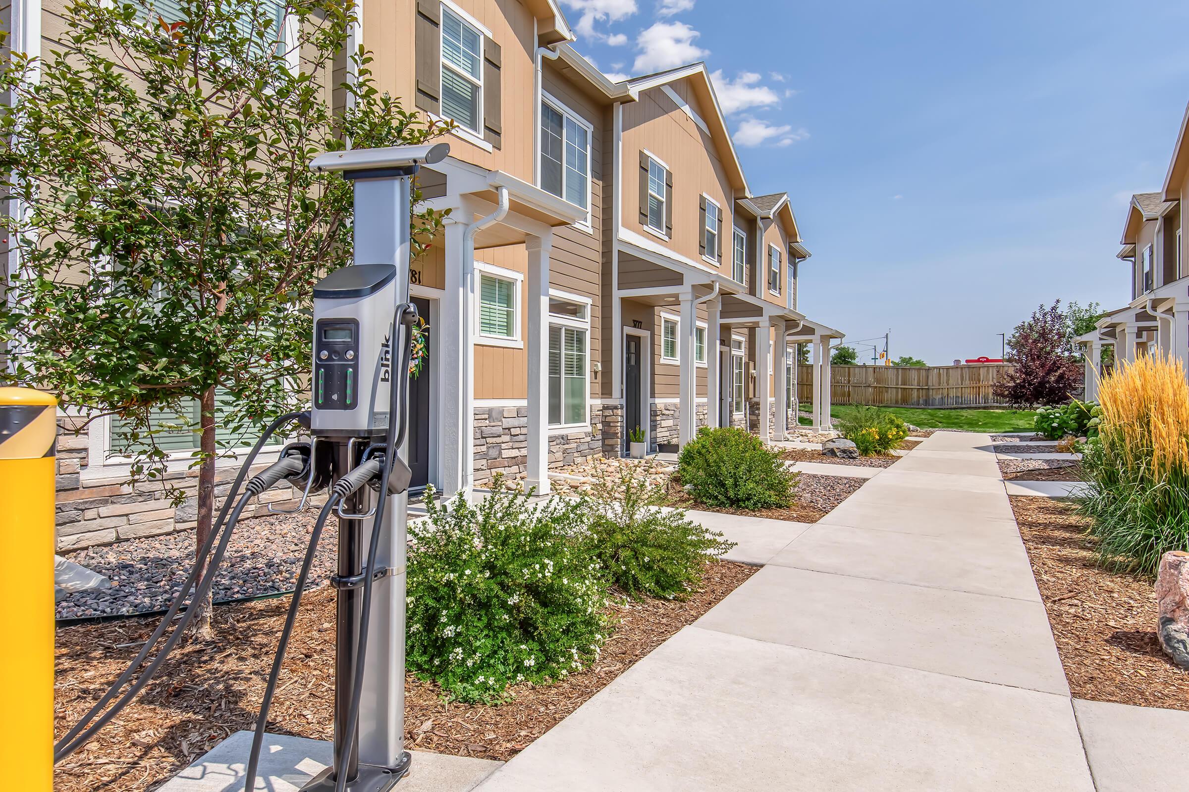 Electric vehicle charging station located in front of residential buildings. The pathway is lined with landscaping, including shrubs and ornamental grasses, creating a welcoming environment. Clear blue sky overhead and vibrant landscaping enhance the scene.