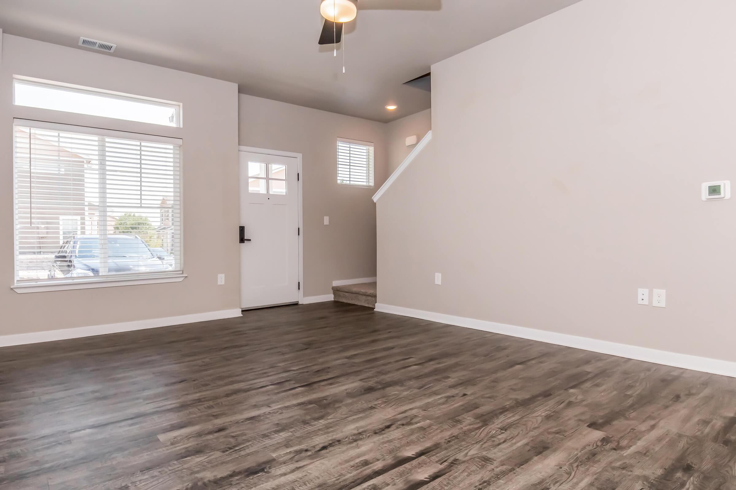 A spacious, empty living room featuring light-colored walls, large windows with blinds, and a ceiling fan. The flooring is dark wood, and there is a staircase leading to an upper level. A front door is visible, providing natural light and a view of the outdoors.