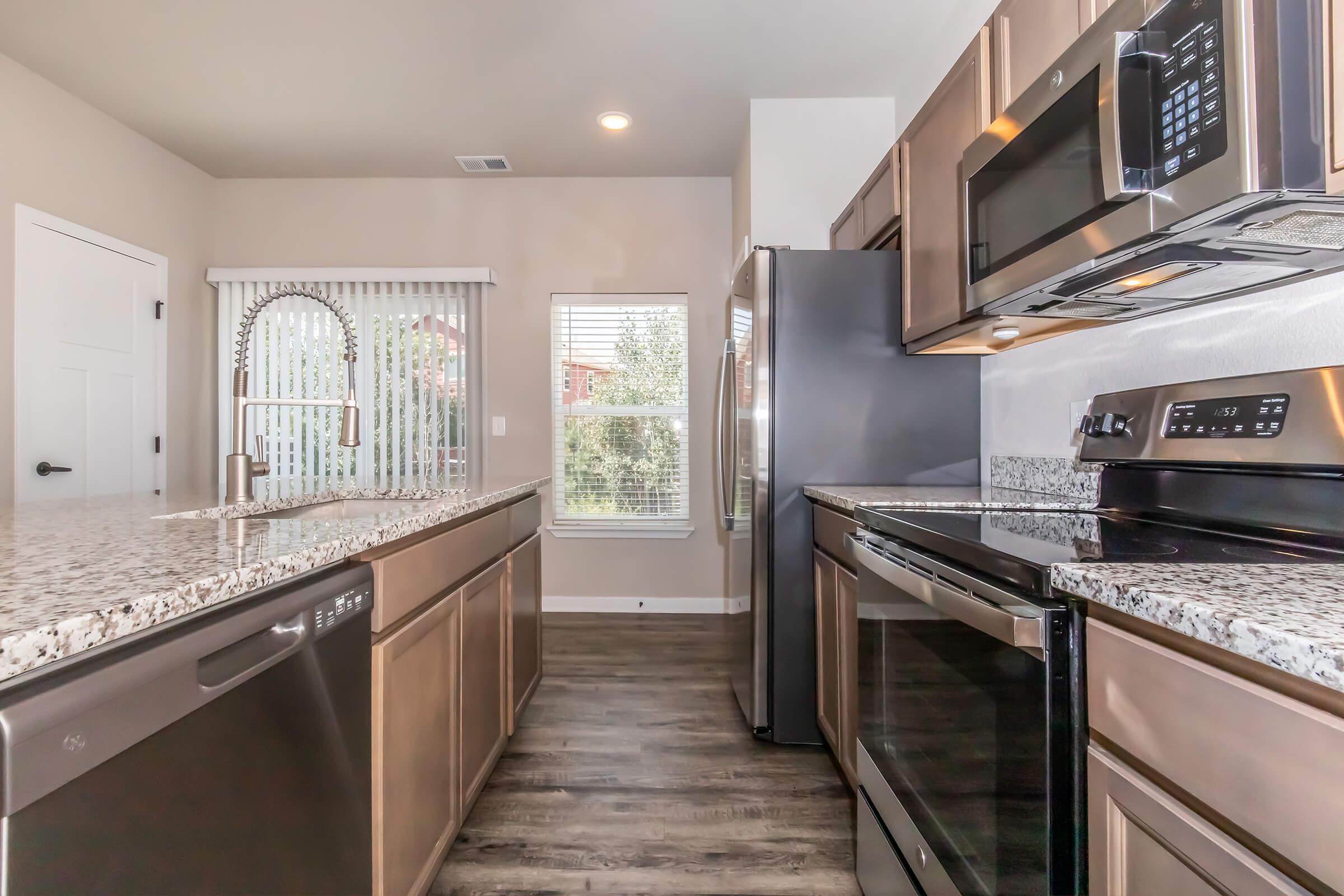 Modern kitchen featuring granite countertops, stainless steel appliances including a microwave and oven, and a dishwasher. The cabinetry is a warm wood tone. A window provides natural light, and the floor has a wood-like finish. Overall, the space is clean and contemporary.