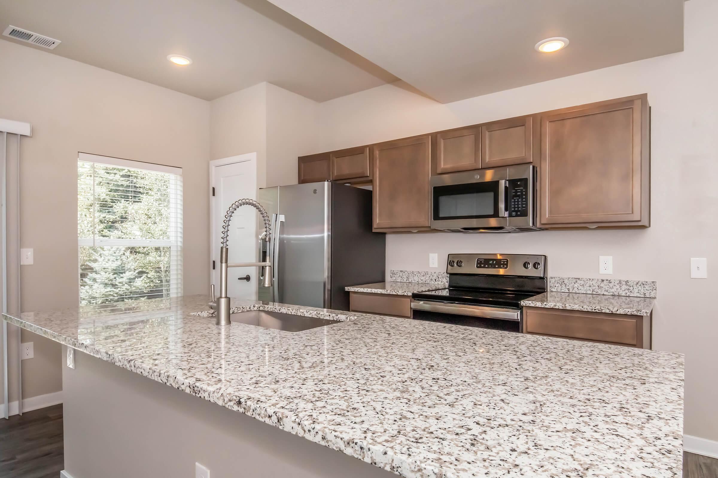 Modern kitchen featuring a large granite countertop with a sink, stainless steel appliances including a microwave and oven, and wooden cabinetry. A window allows natural light to illuminate the space, which is designed with neutral colors for a clean and contemporary look.