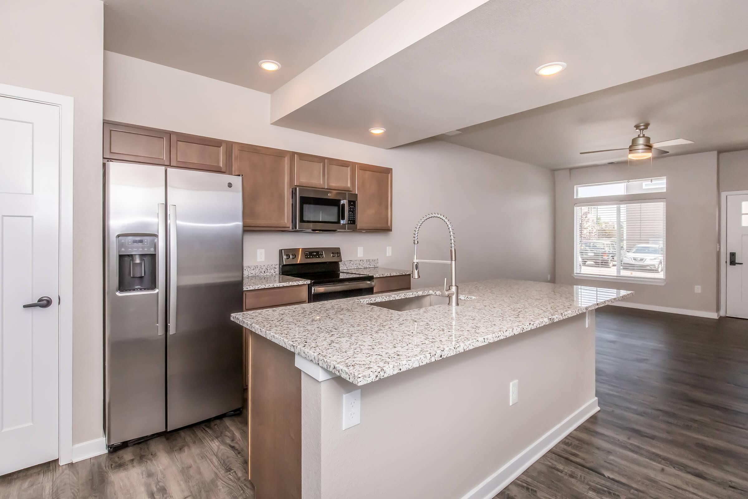 A modern kitchen featuring stainless steel appliances, including a refrigerator and microwave, with wooden cabinetry and granite countertops. The kitchen island includes a sink and is open to a bright living area with large windows, showcasing a contemporary design.