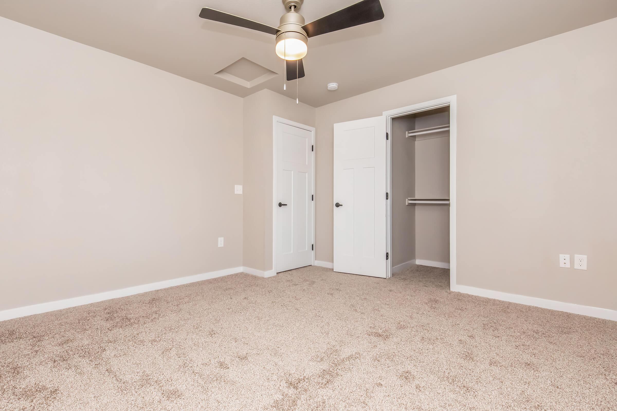 A spacious room with light beige walls and plush carpet. The room features two white doors, one leading to a closet, and has a ceiling fan in the center. Natural light appears to illuminate the space, creating a warm and inviting atmosphere.