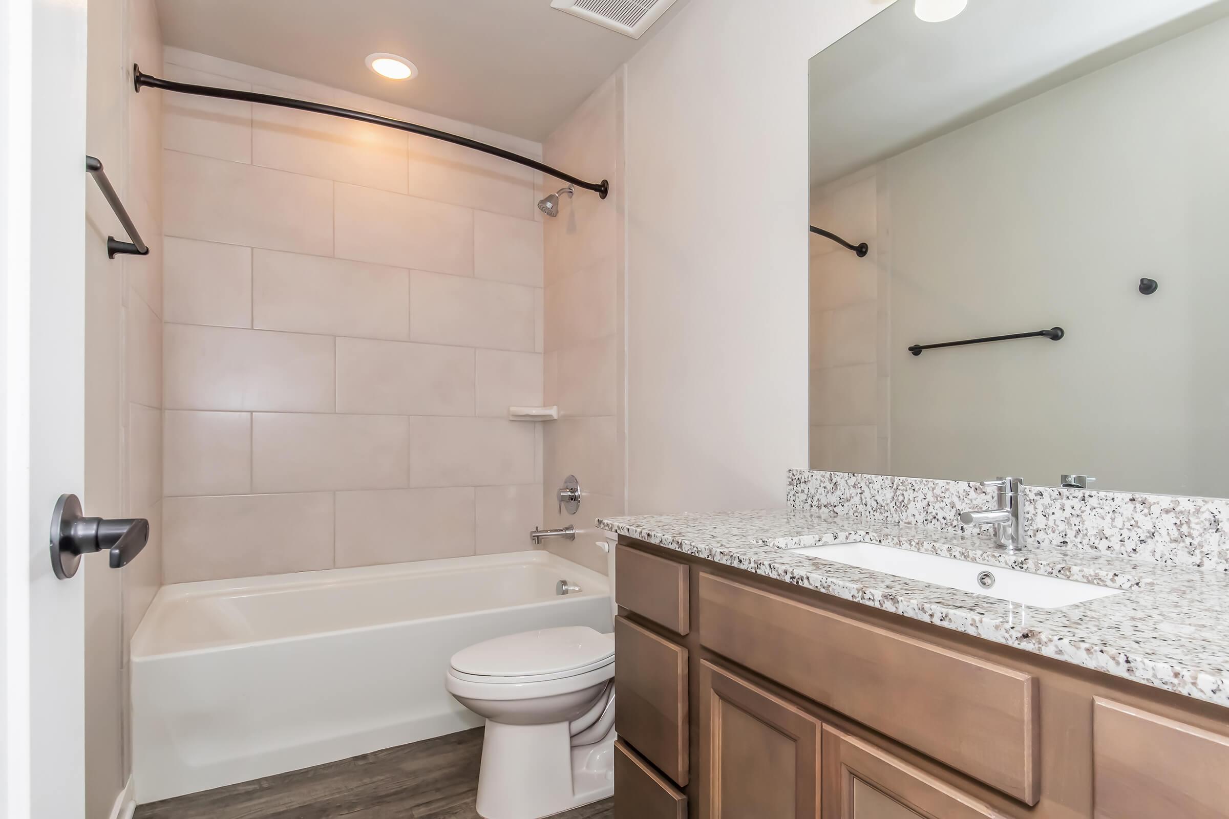 A modern bathroom featuring a white bathtub and shower area, a granite countertop with a sink, wooden cabinetry, and a large mirror. The walls are lined with light-colored tiles, and there is overhead lighting. A towel bar is mounted on the wall, and the floor has a wood-like appearance.
