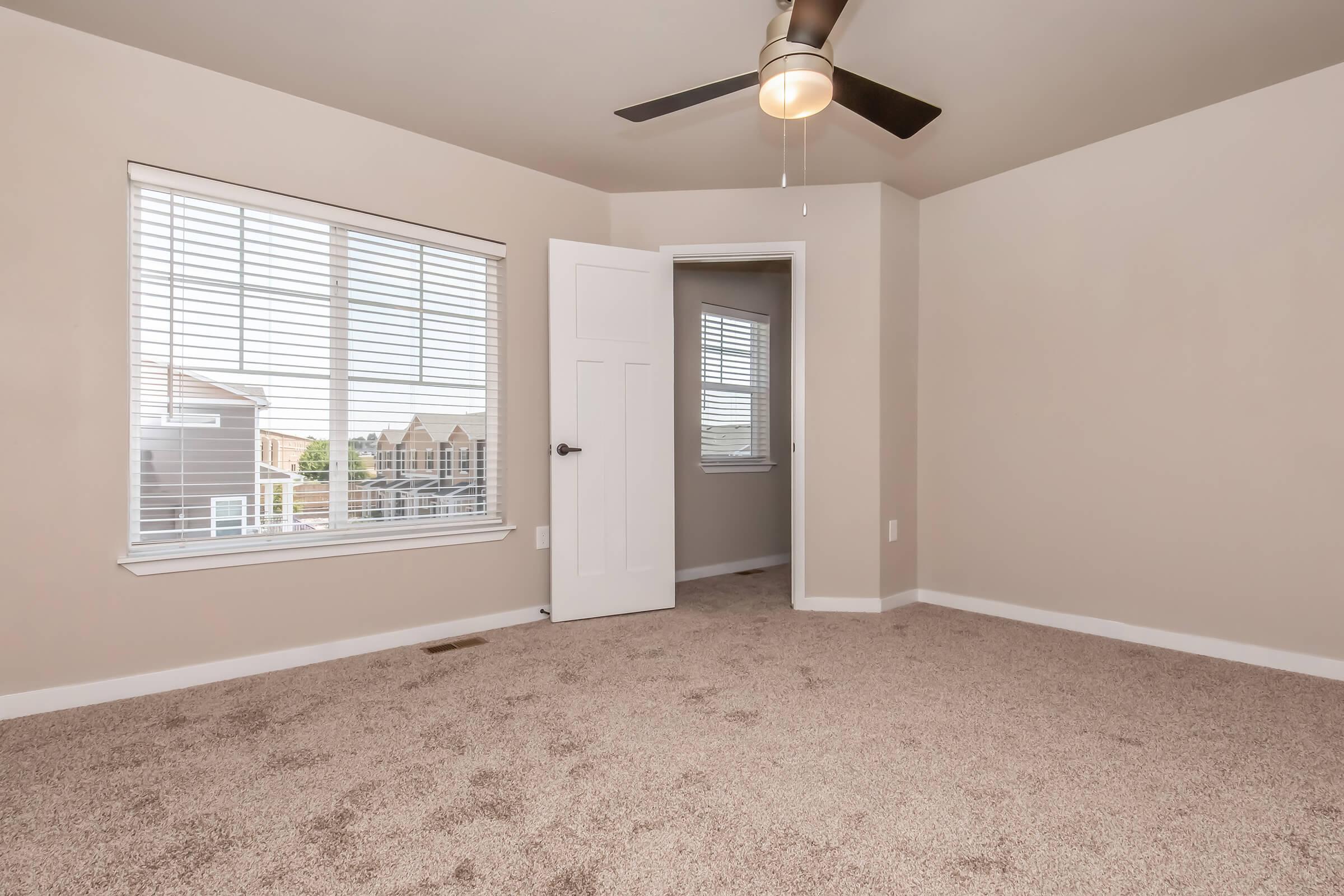 A well-lit bedroom featuring beige walls, a ceiling fan, and carpeted flooring. There is a large window with blinds allowing natural light to enter, and an open door leading to another room. The space is uncluttered and ready for furniture placement.
