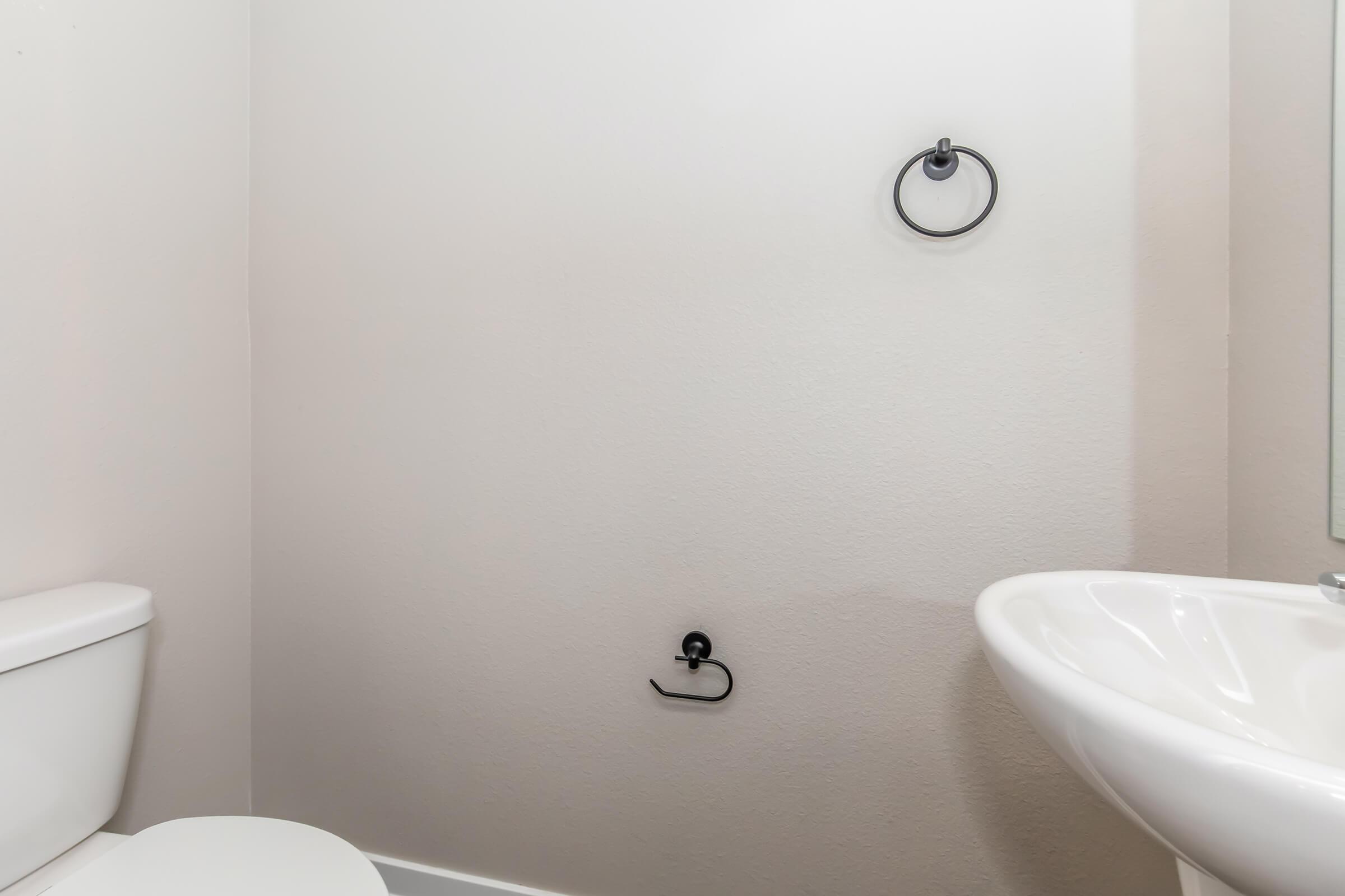 A bathroom interior featuring a white toilet and a small sink. The wall is plain and light-colored, with a towel ring mounted on one side and a black towel holder visible below it. A mirror reflects the light, contributing to a clean and simple design.