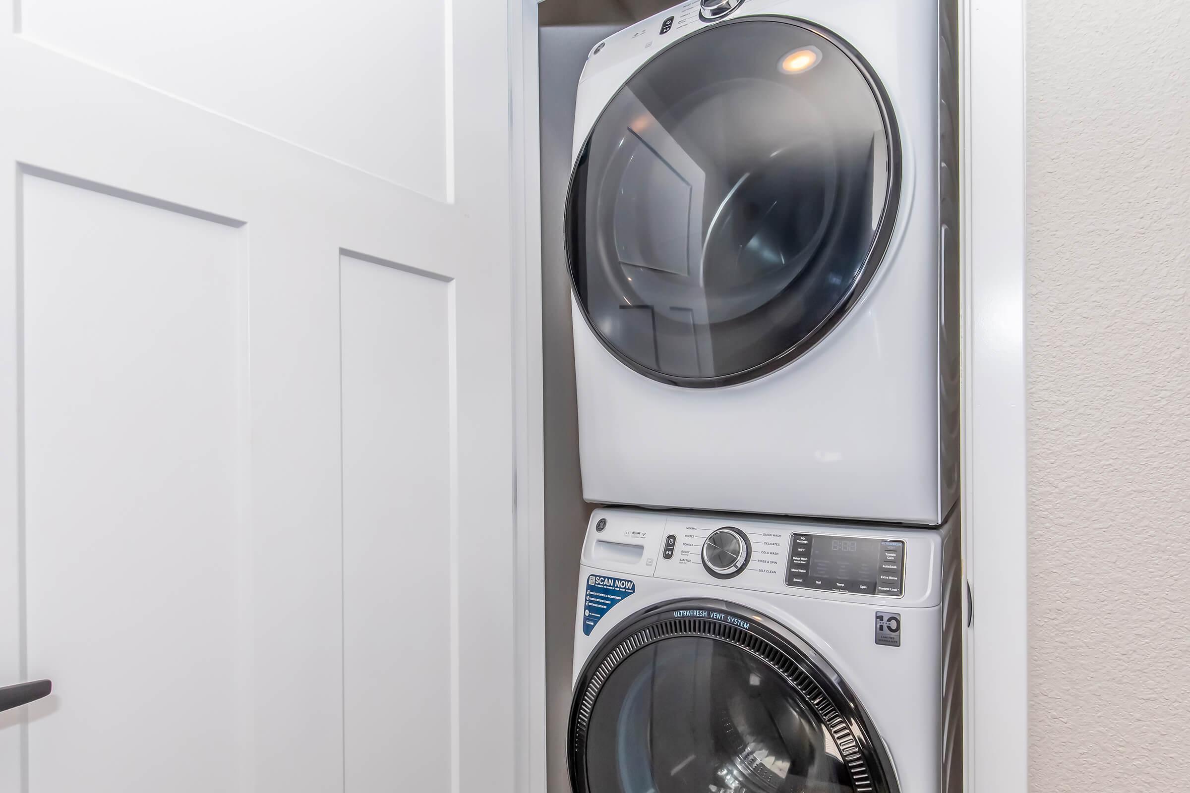 A stacked washer and dryer in a compact laundry space with a white door. The appliances are modern, featuring glass doors and a control panel, set against a light-colored wall.