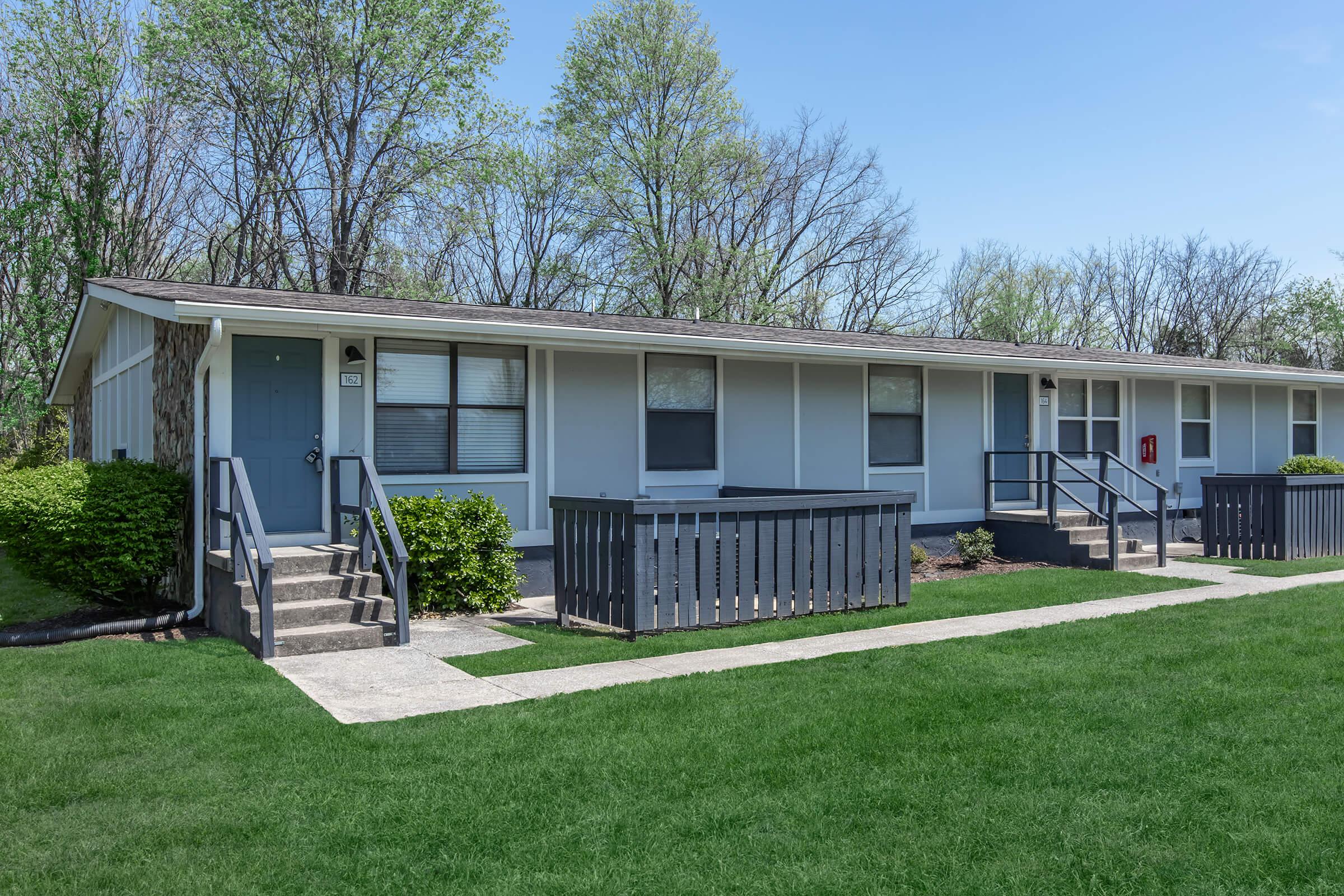 A row of single-story apartment buildings with a light blue exterior, surrounded by well-manicured green grass and small shrubs. Each unit has a front door with steps leading up, and there are railings along the pathways. Trees are visible in the background under a clear blue sky.