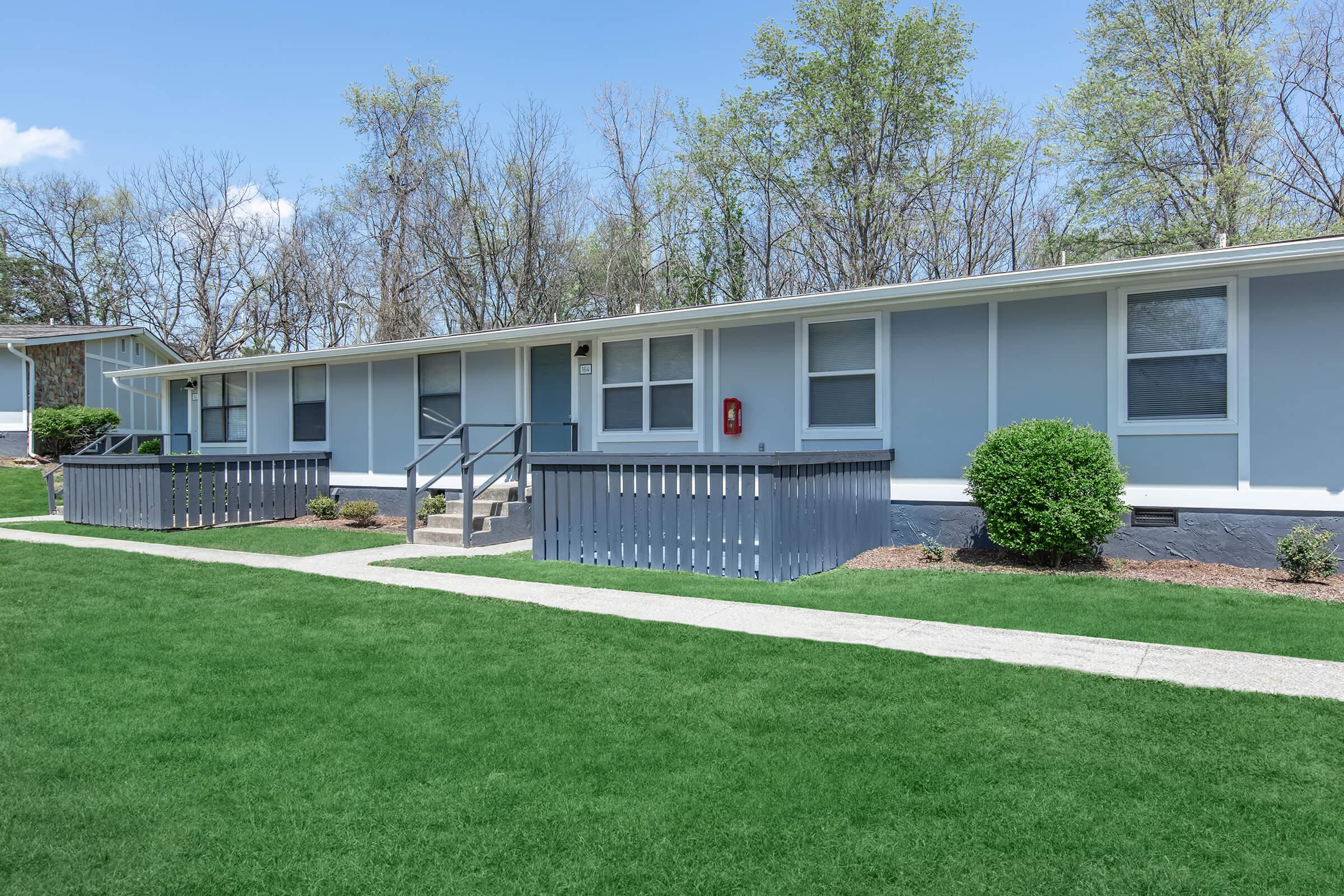 A modern single-story building with gray siding and large windows, surrounded by a well-maintained lawn. The building features a walkway leading to the entrance, along with gray railings on the porch. Trees in the background indicate a natural setting, and the sky is clear blue.