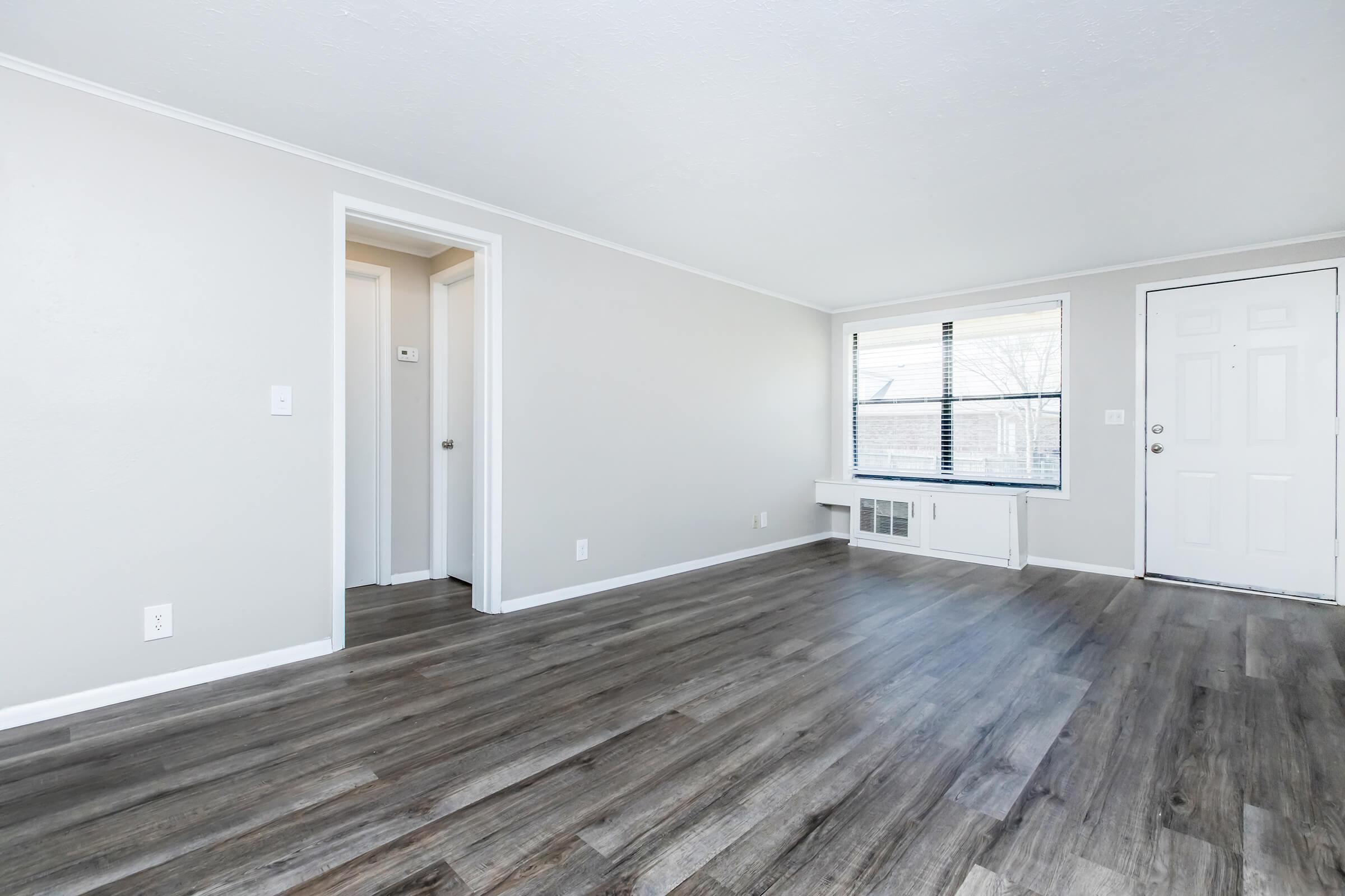 A spacious, empty living room with light gray walls and brown laminate flooring. There are two doorways, one leading to a hallway and the other to the front entrance, which has a window beside it. Natural light enters through the window, illuminating the room.