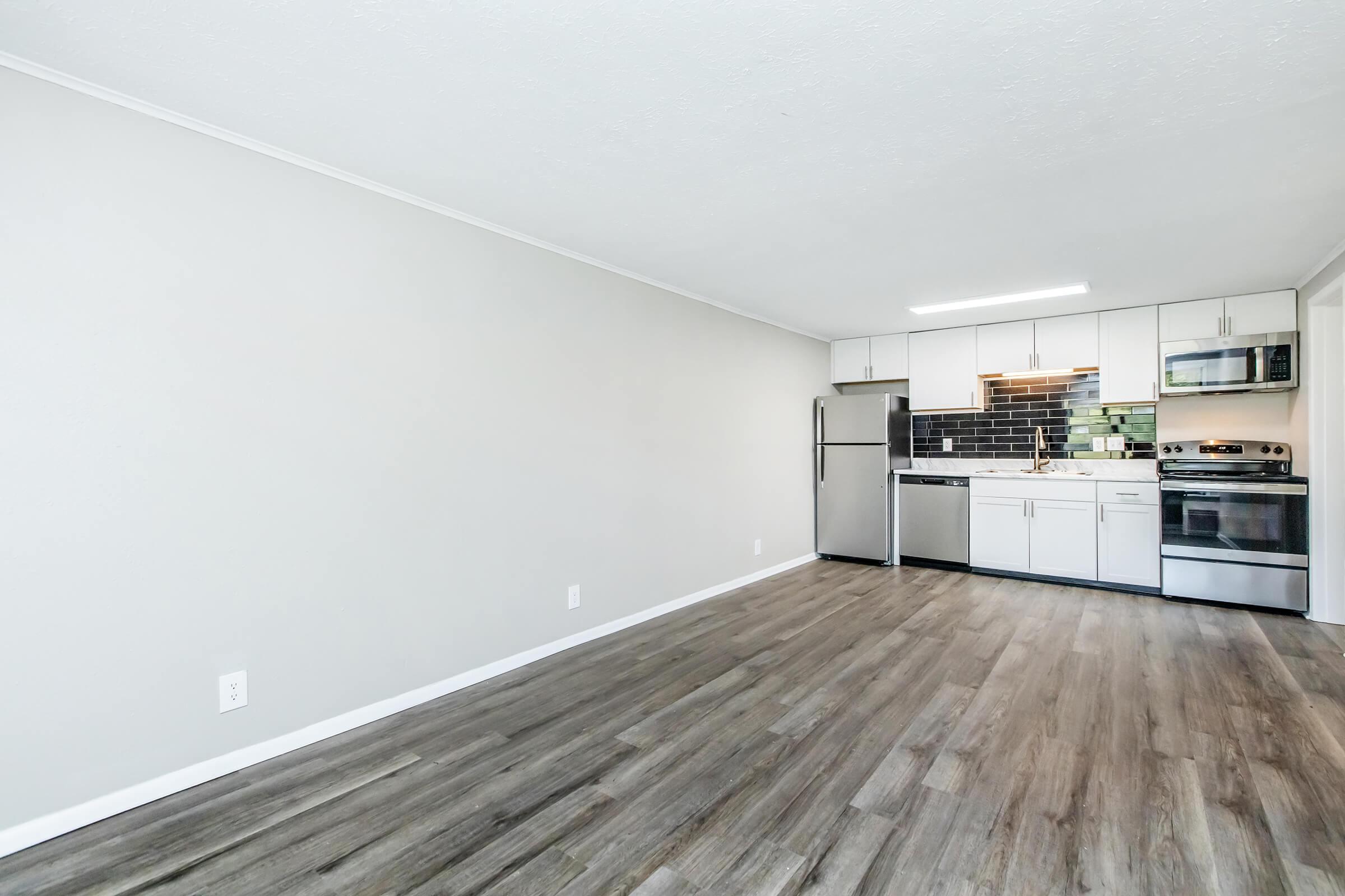 A modern kitchen and living area featuring gray walls, a stainless steel refrigerator, oven, and microwave, with dark tiled backsplash. The floor is wood laminate, and there is ample natural light from a nearby window. The space is minimally decorated, creating a clean and open feel.
