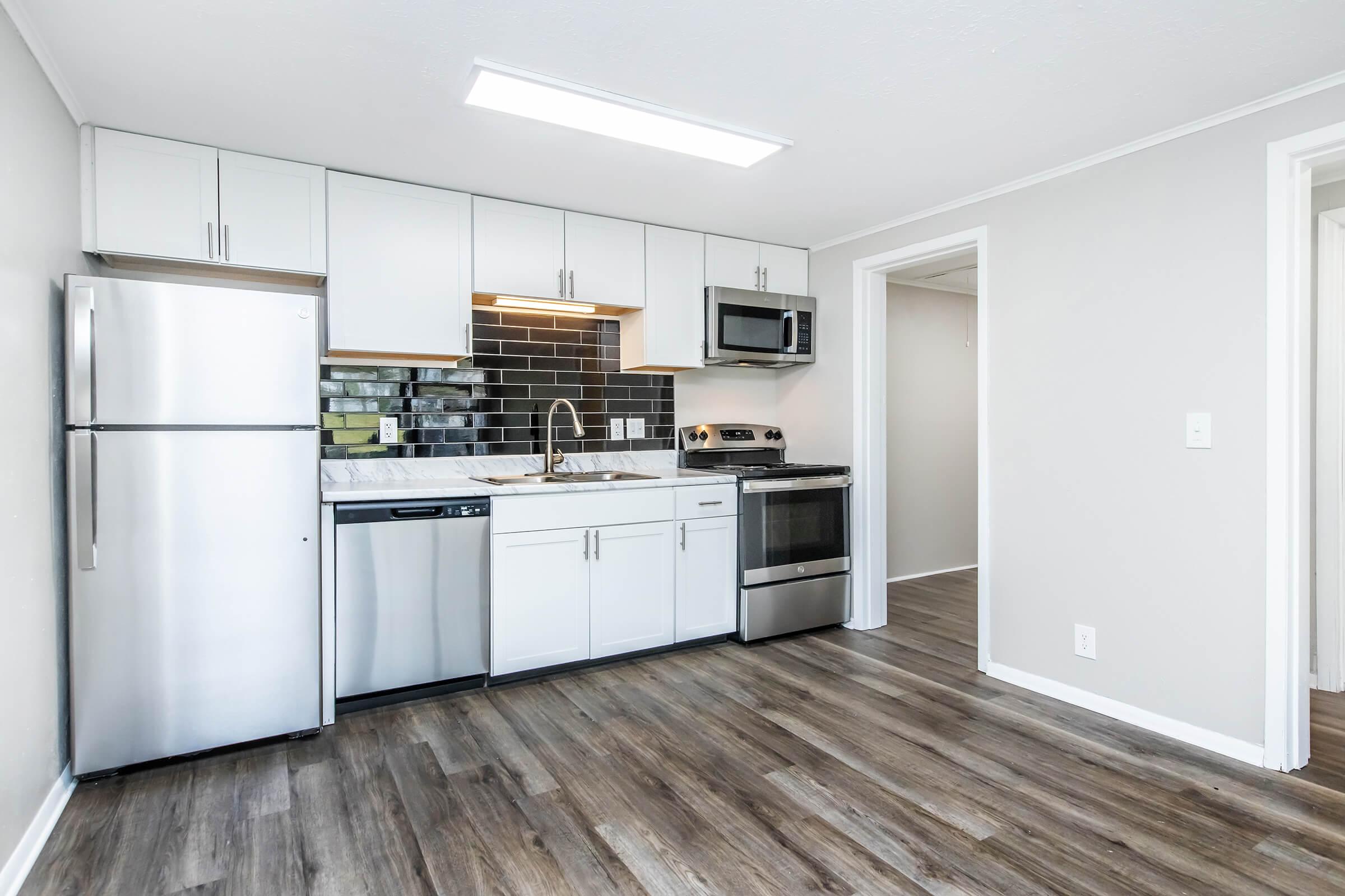 Modern kitchen featuring stainless steel appliances, including a refrigerator, dishwasher, and oven. White cabinets and a black tiled backsplash provide a sleek look. The space has laminate flooring and a bright overhead light, creating an inviting atmosphere. An open layout leads to an adjacent room.