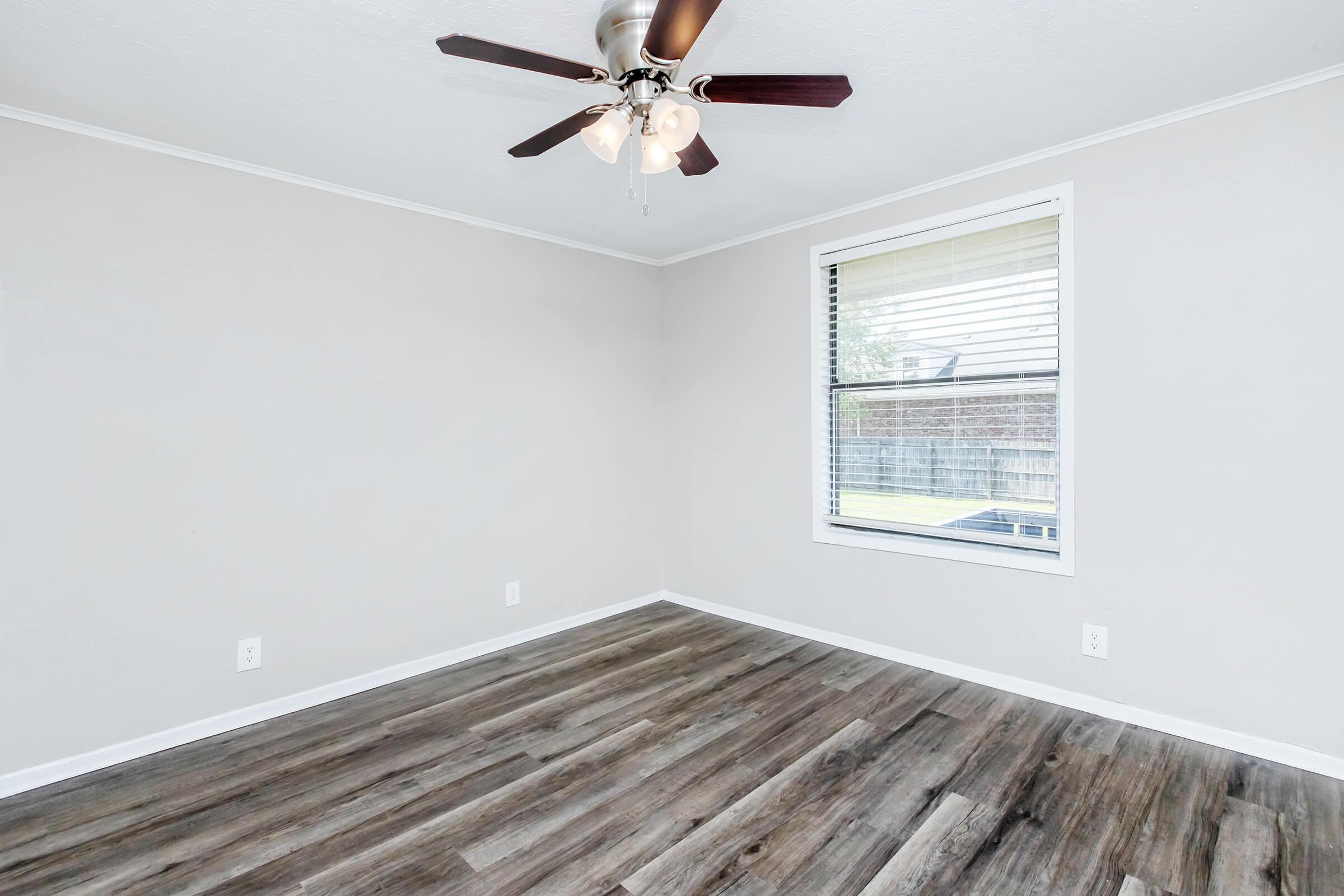 A spacious, empty room with light gray walls and a ceiling fan. The floor features wood-like laminate, and a window with blinds allows natural light to enter, revealing a backyard view.