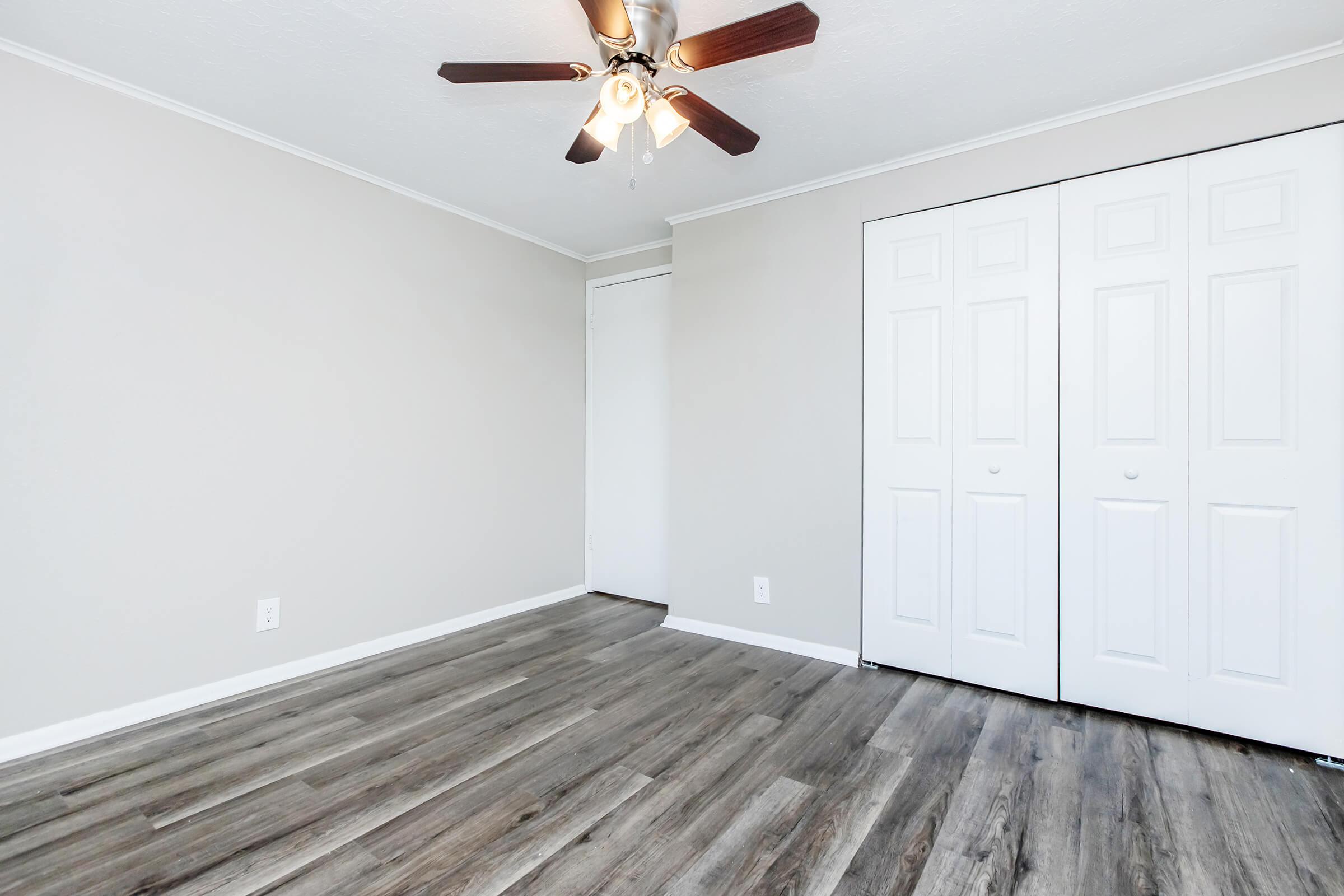 A clean, empty room with light gray walls and a ceiling fan. The floor features light-colored laminate wood. There are white sliding closet doors on one wall, and a small door leads to another area, likely a closet or bathroom. The overall appearance is simple and modern.