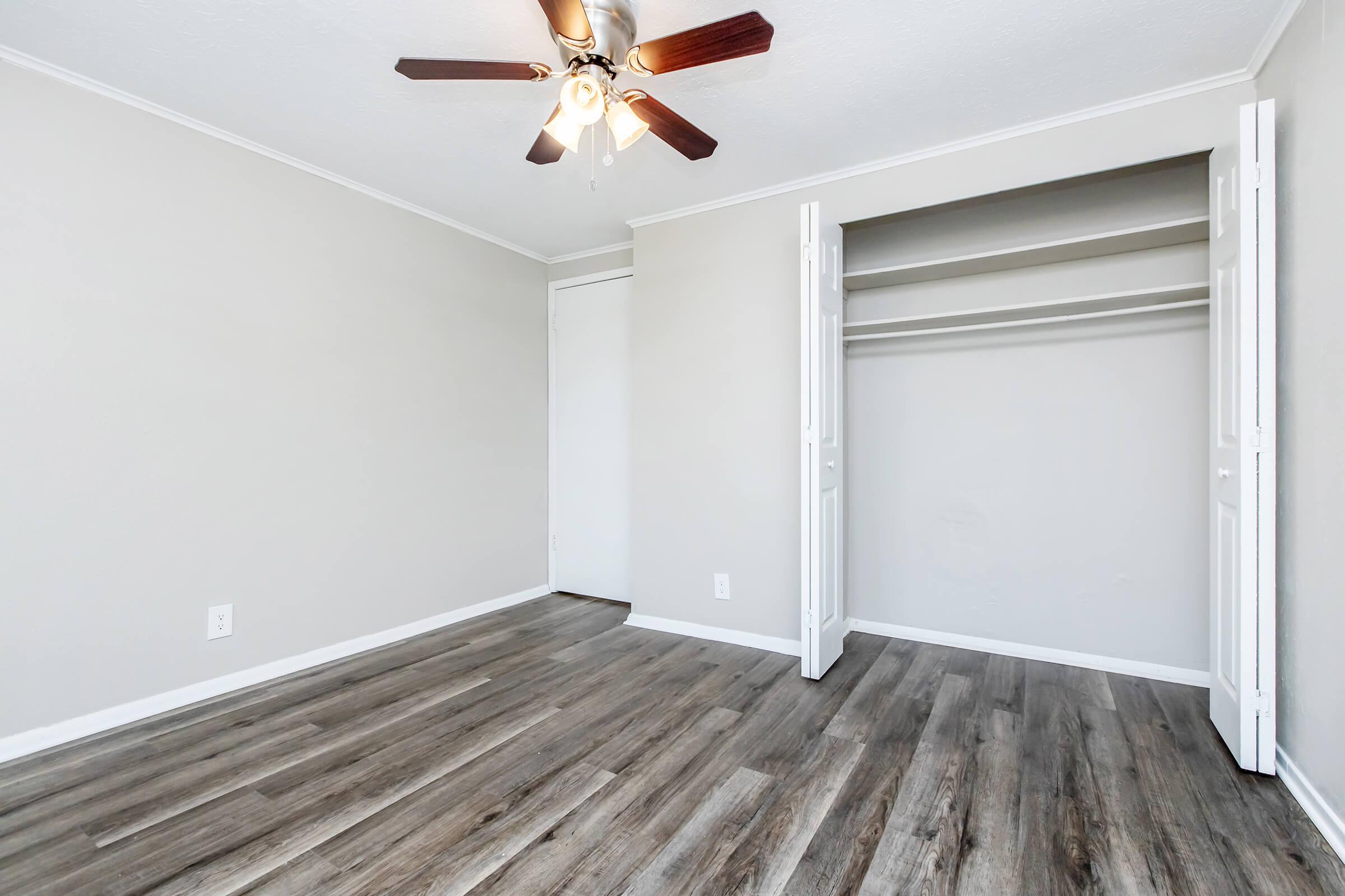 A well-lit, empty room with light gray walls and a ceiling fan. The flooring is a modern, light wood style. There is a closet with open double doors on one side, showcasing an empty space. The overall appearance is clean and ready for new furnishings.