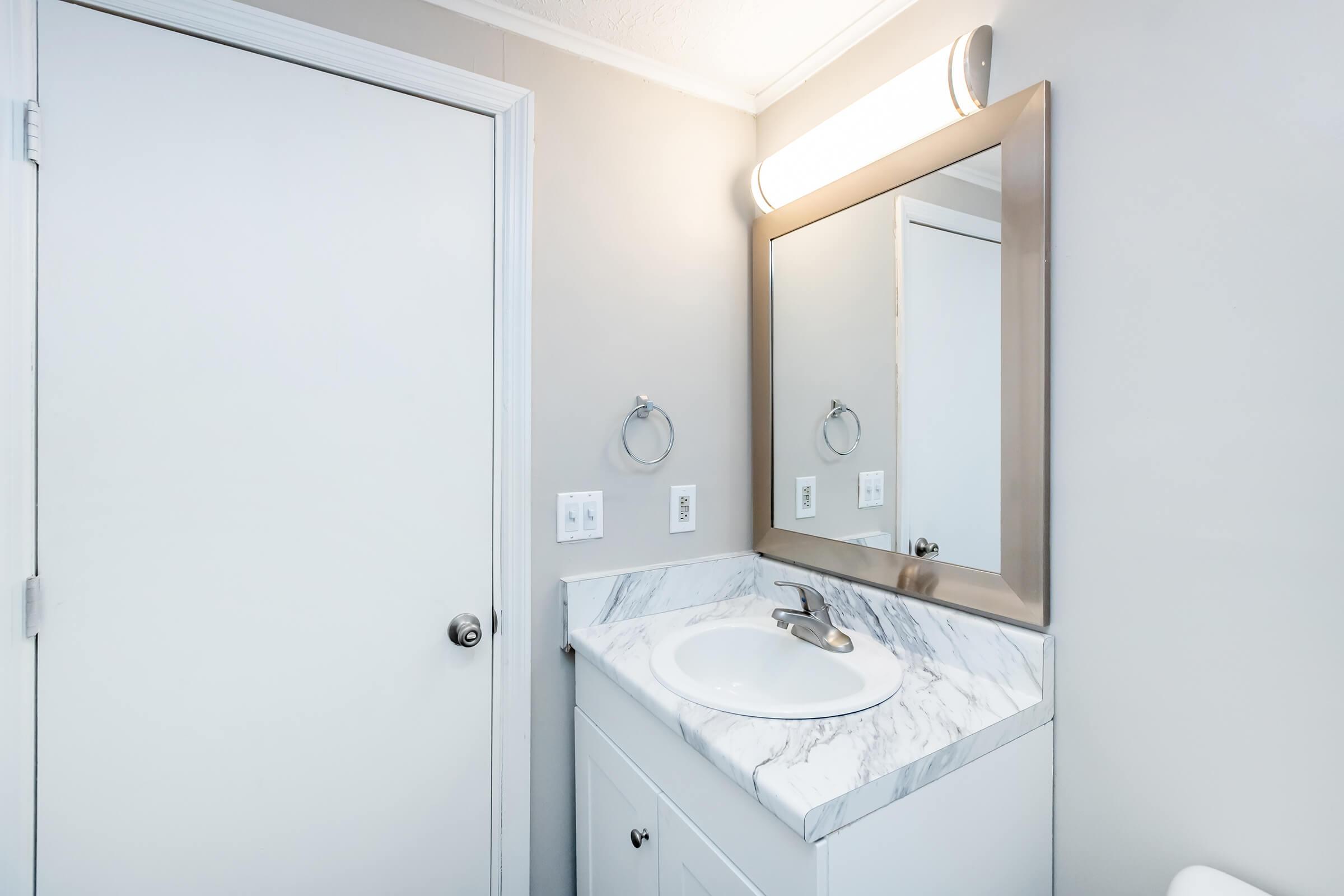 A well-lit bathroom featuring a white vanity with a marble countertop, a modern mirror with a metallic frame, and a simple faucet. The walls are painted a light gray, and there is a white door and towel holder visible in the background. The space is tidy and contemporary.